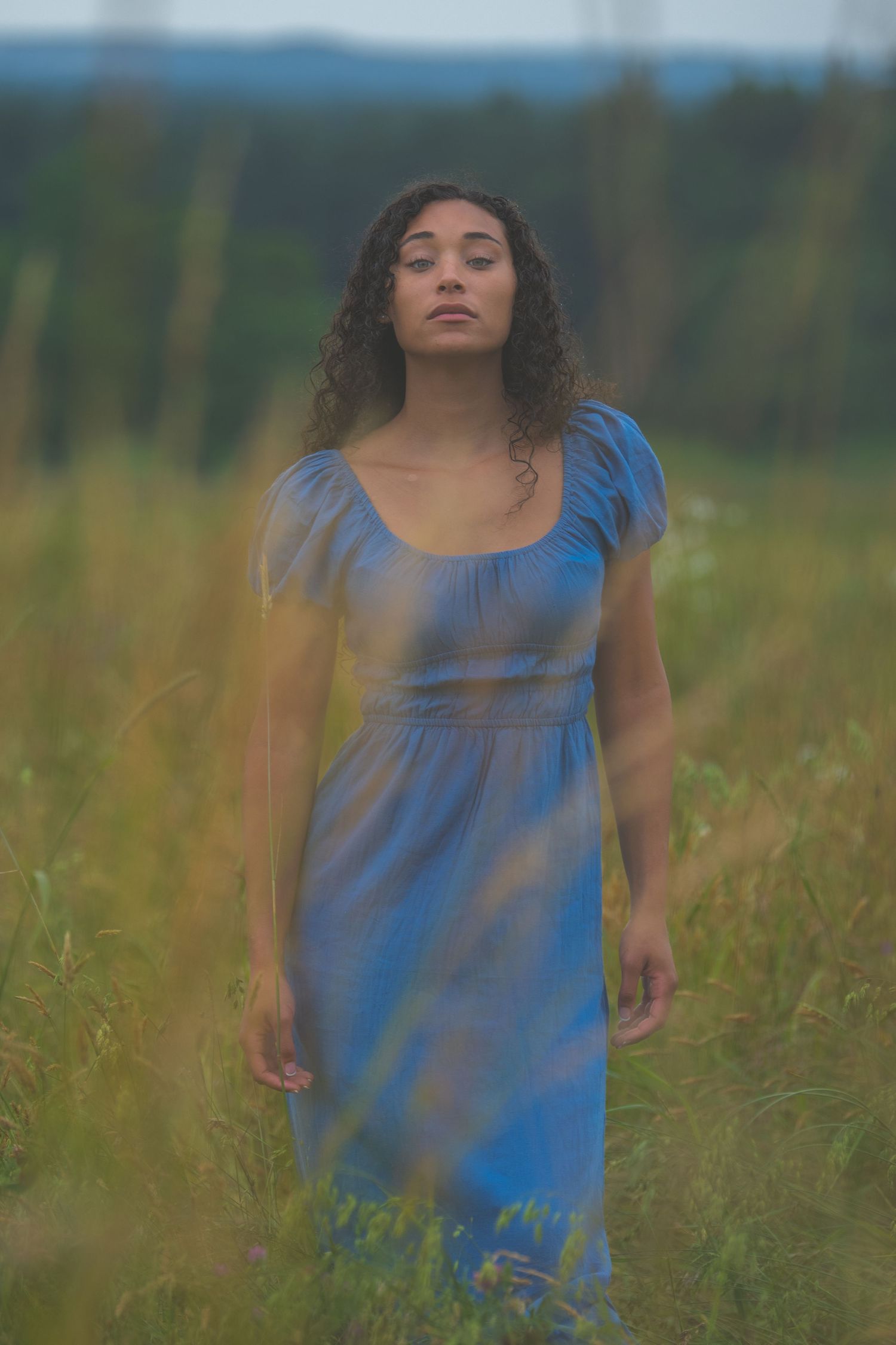 Woman in blue dress stands serenely among tall grass at sunset.