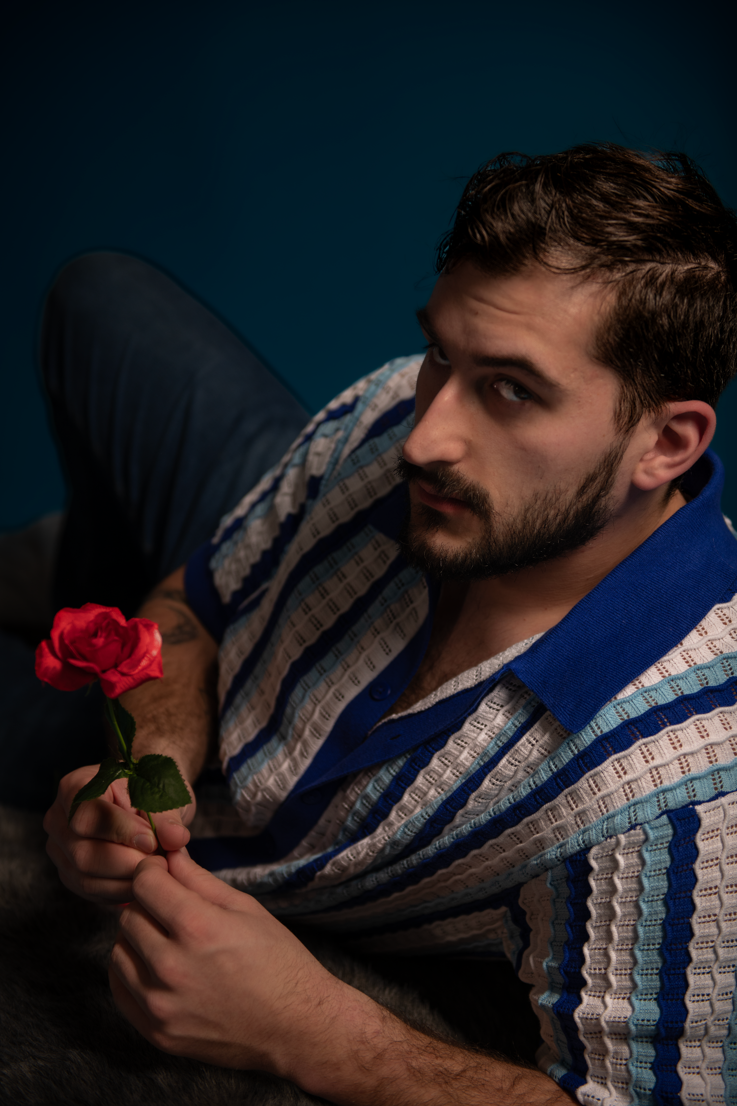A man in a plaid shirt and dark jacket poses with a red rose in a dramatic studio lighting setup.
