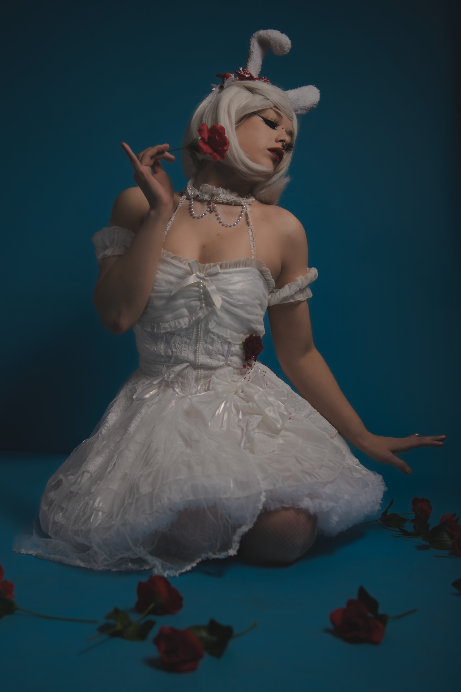A person in a white lacy dress and bunny ears poses elegantly with red roses scattered on a blue background.