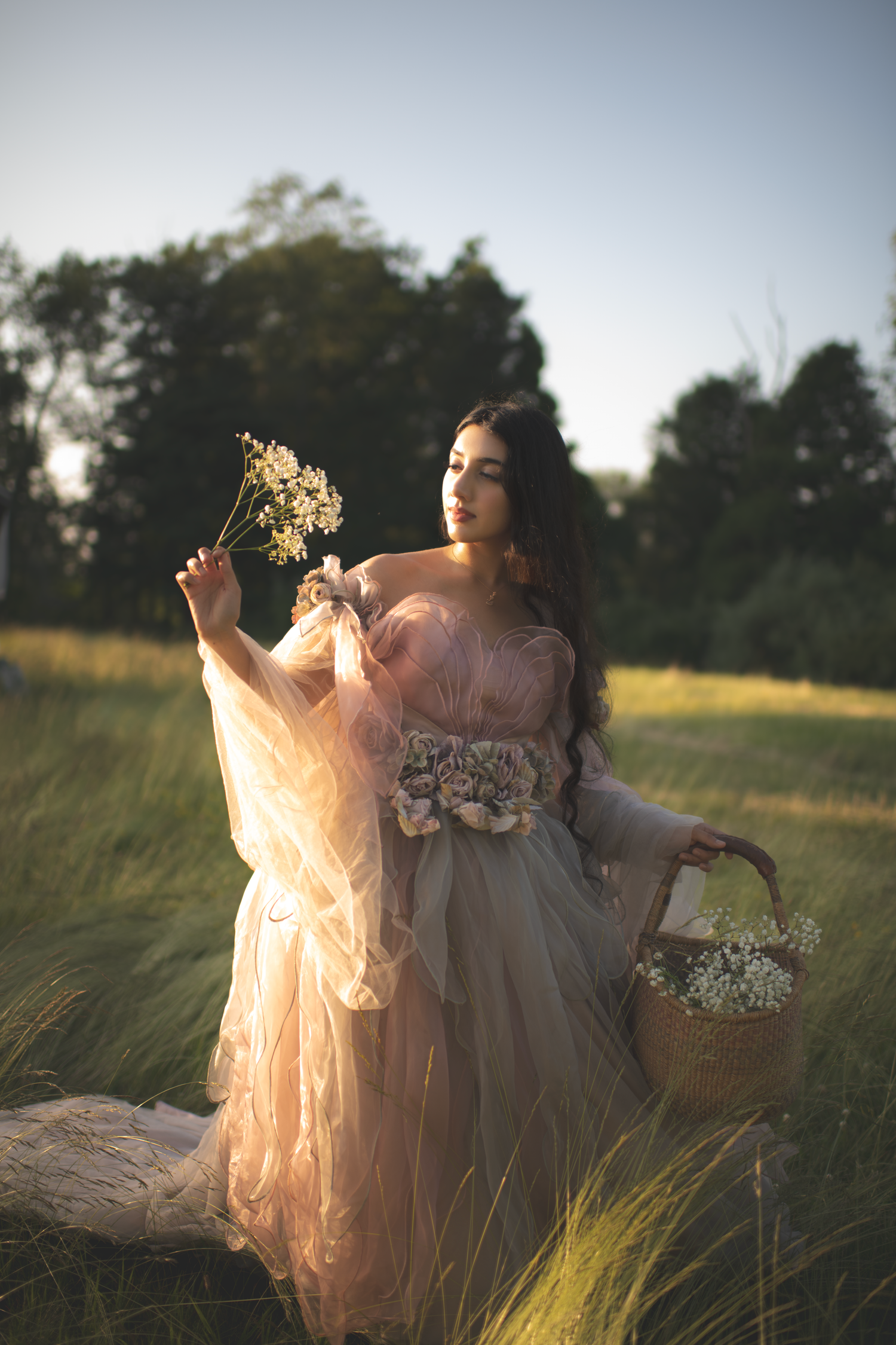 Romantic outdoor photoshoot featuring flowing pink gown and delicate flowers against a natural meadow backdrop.