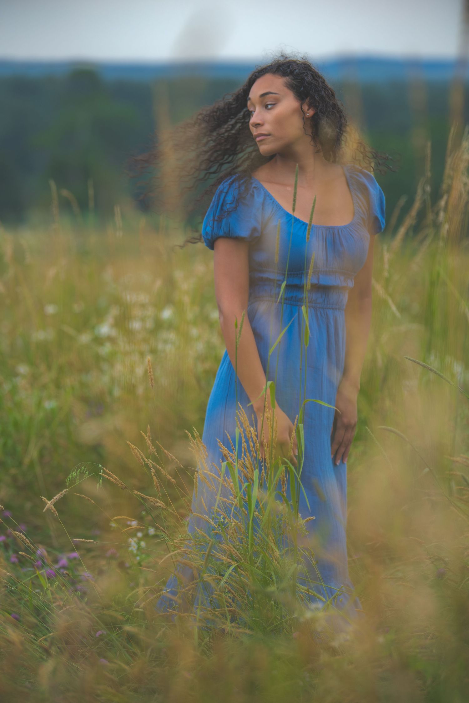Dreamy portrait of a woman in blue dress standing among tall summer grasses.