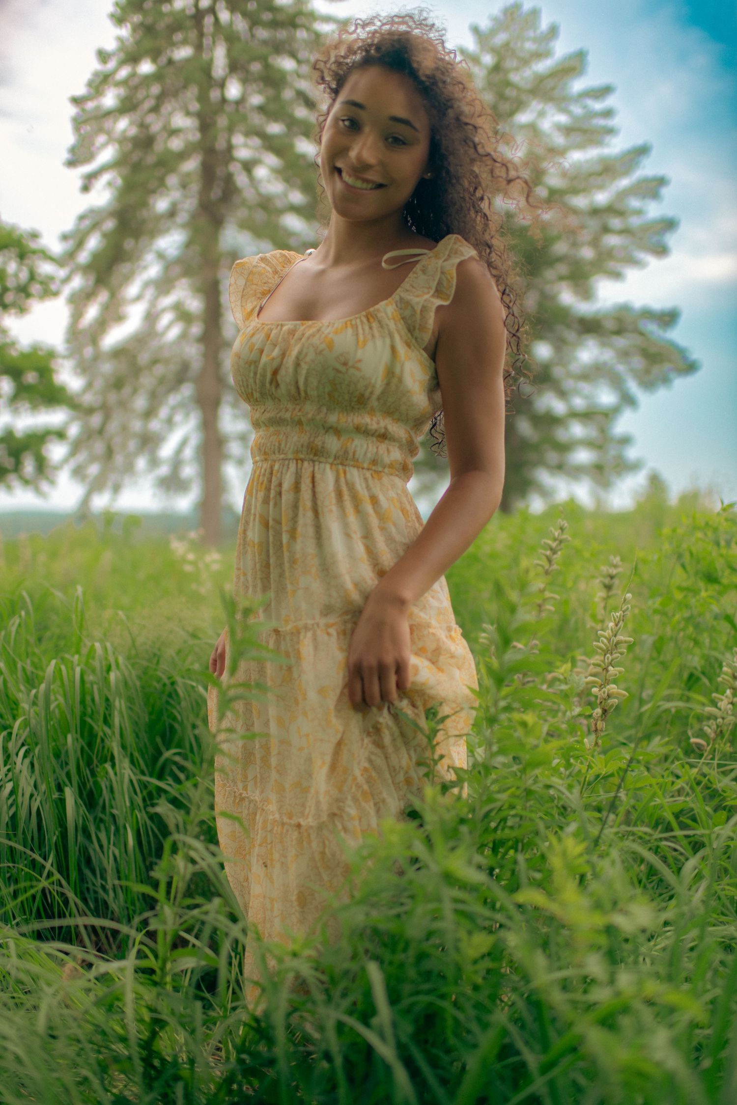 Woman wearing a flowing yellow dress stands among tall grass on a summer day.