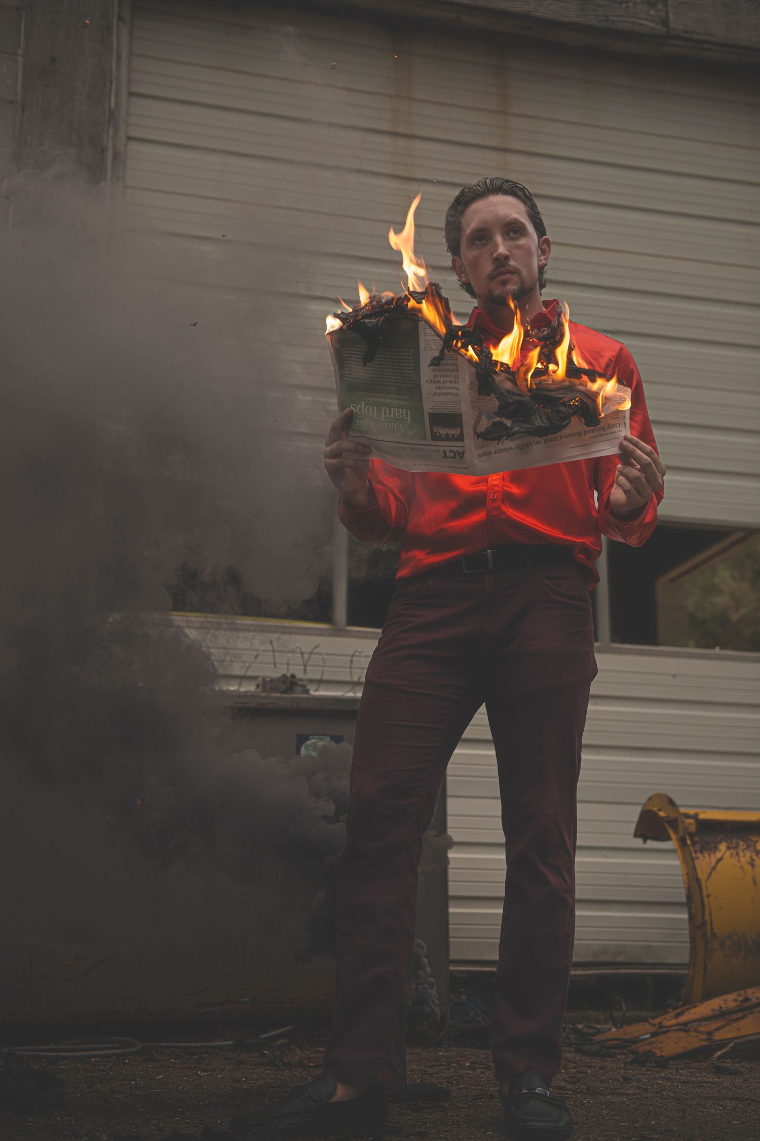 Someone in a red jacket holds a flaming book or magazine while standing outdoors in a smoky setting.