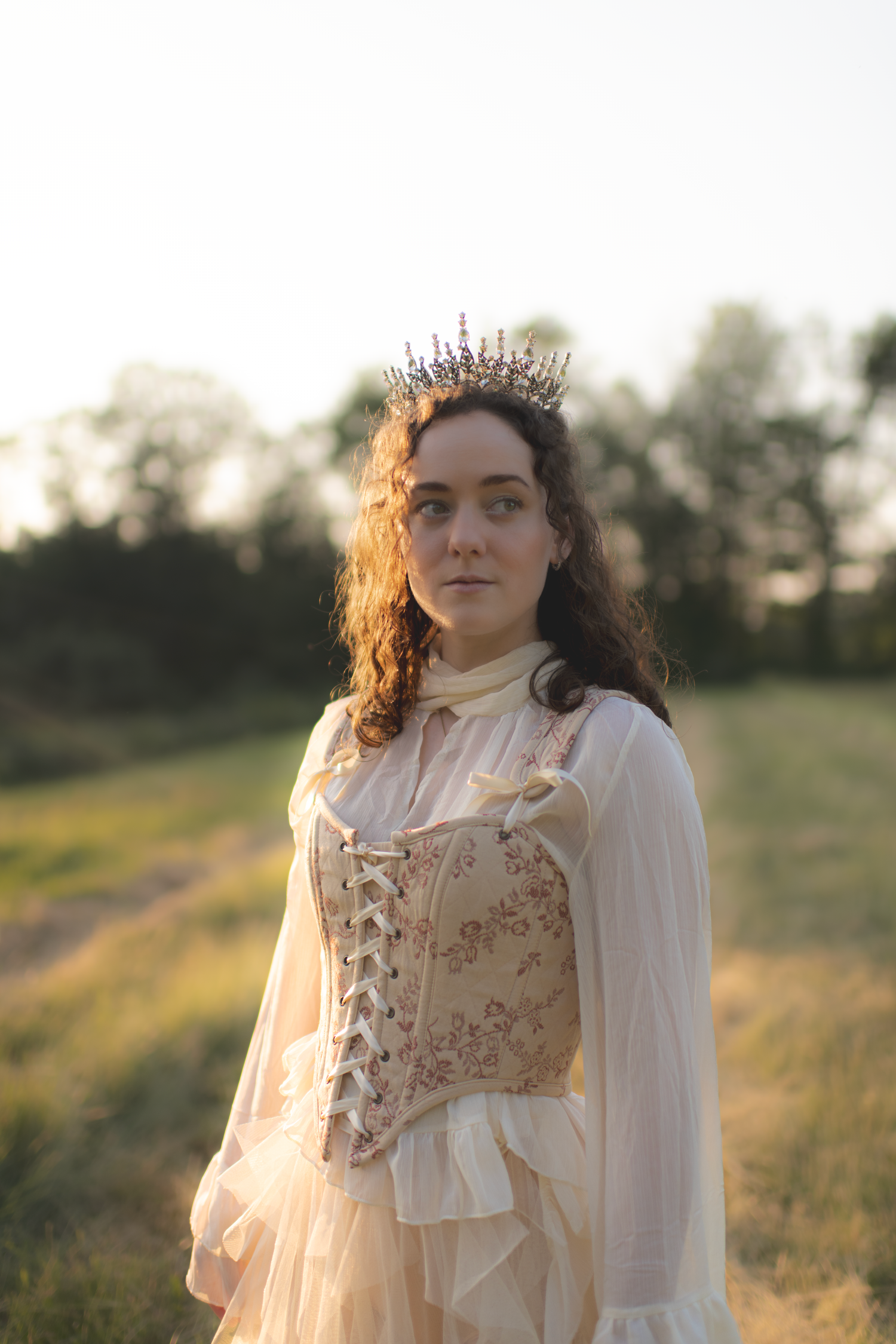 A person in a vintage-inspired white dress and corset poses in a sunlit meadow at golden hour.