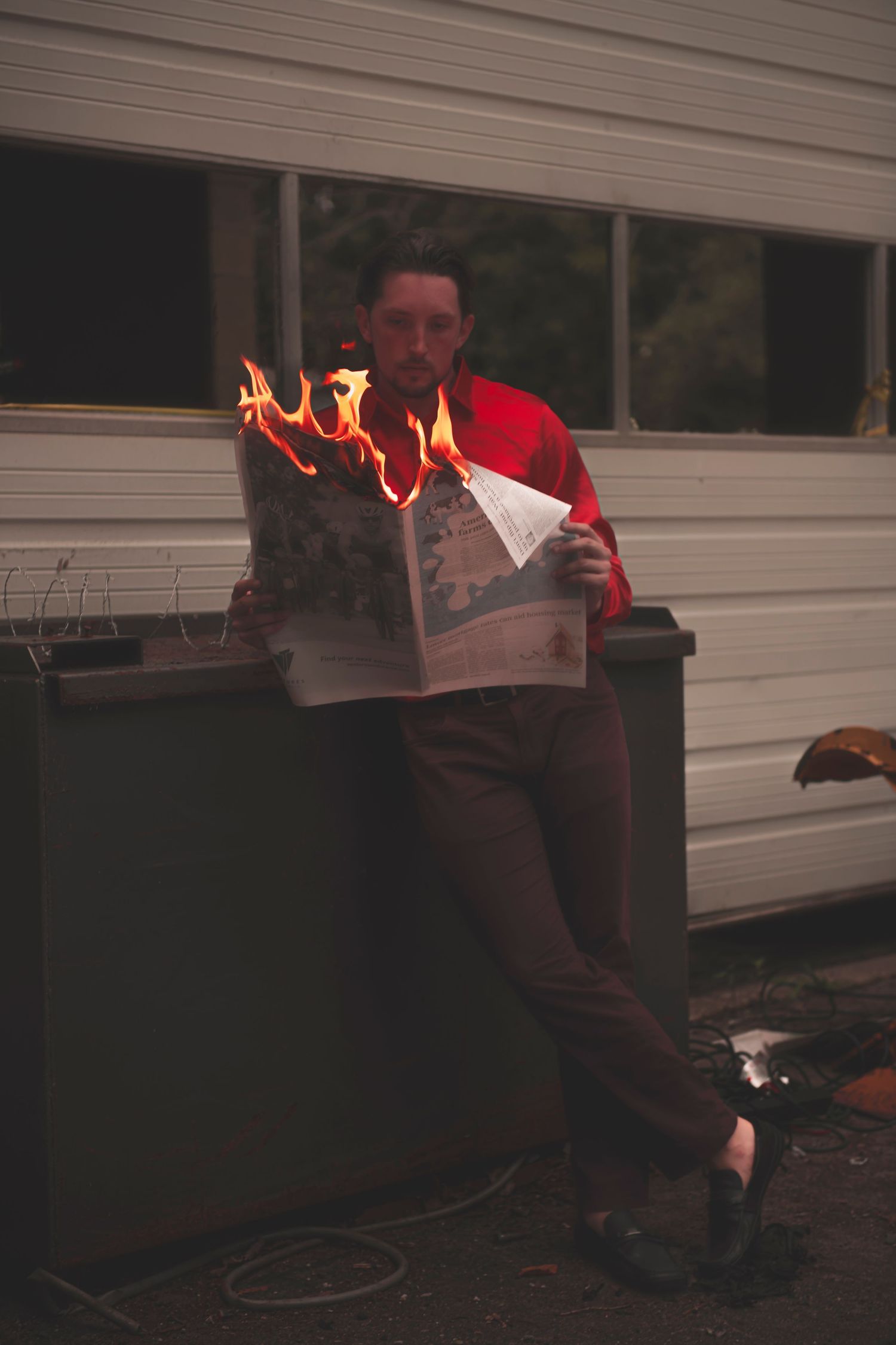Someone in a red sweater leans against a wall while reading a newspaper that appears to be on fire.