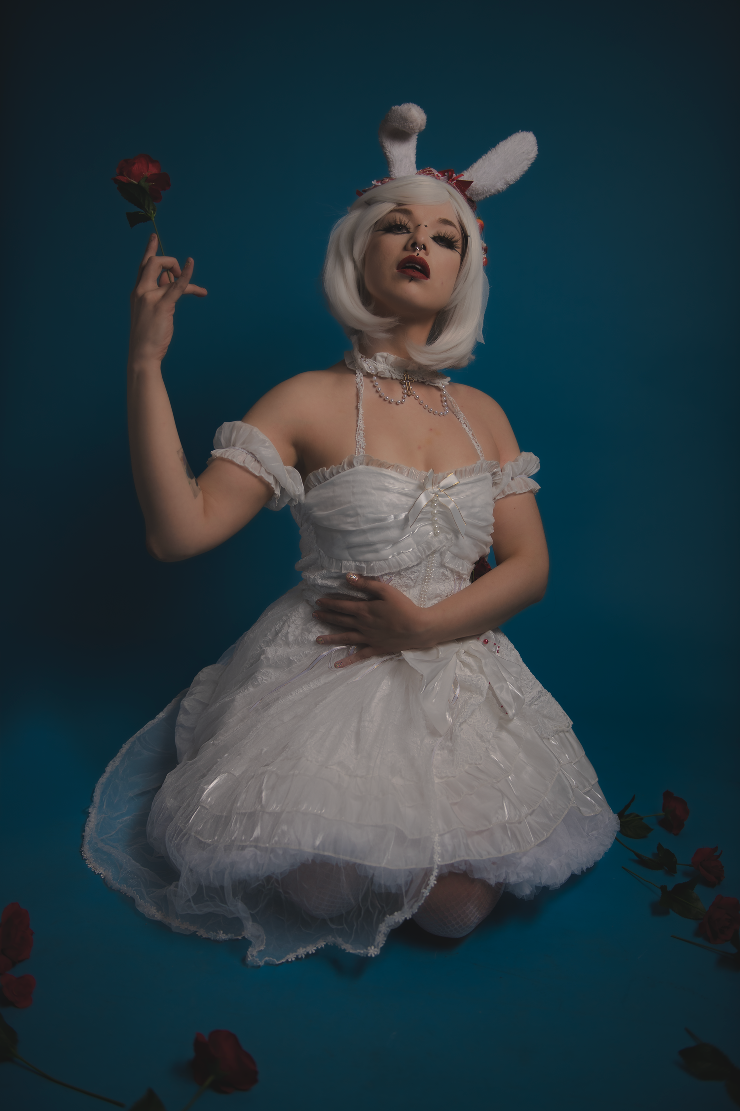 Model in white tulle dress with bunny ears poses on dark blue backdrop with scattered red flowers.