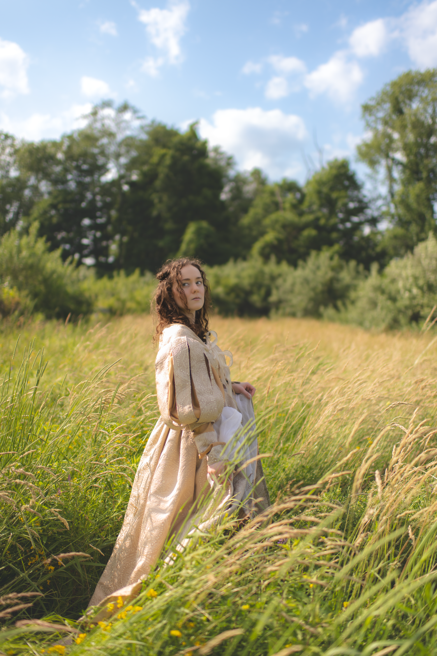 A person in a cream-colored dress walks through tall golden grass in a sunny meadow.