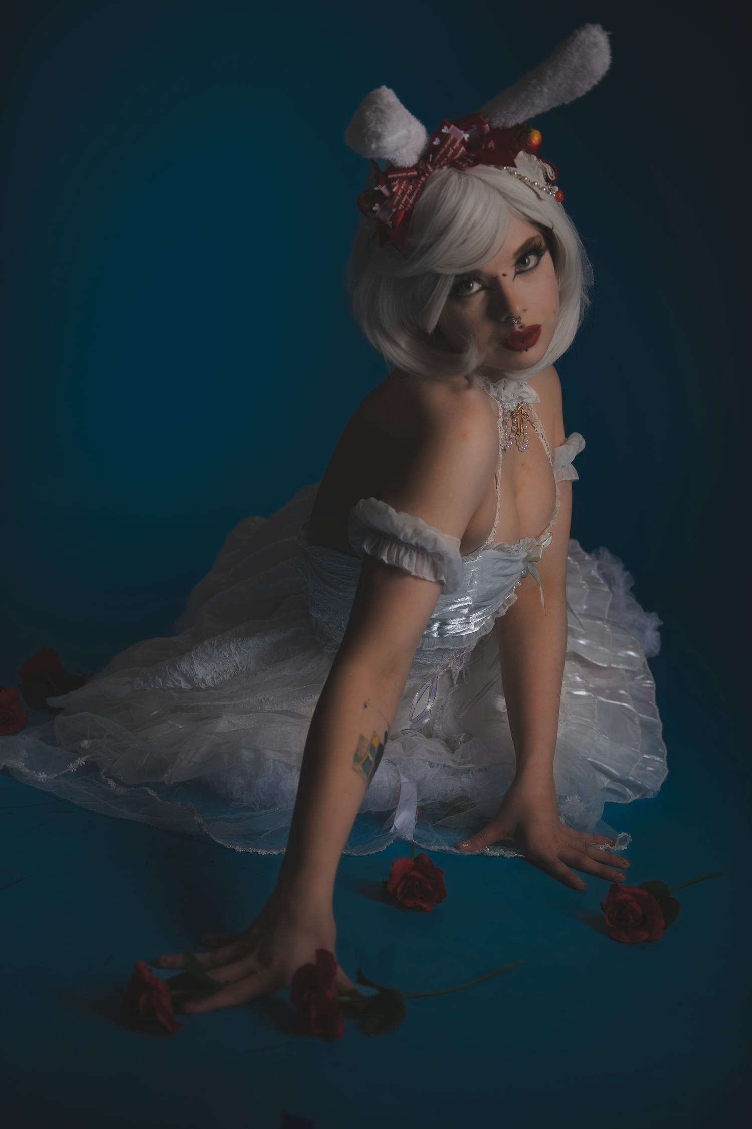 Figure in white dress and bunny ears poses on floor with scattered red petals.