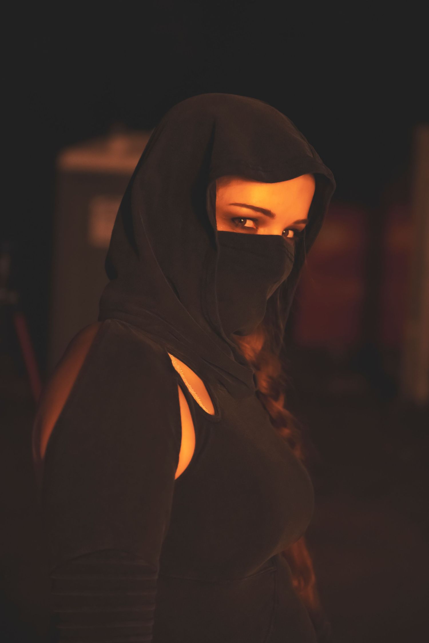 Mysterious figure in black hijab and face covering illuminated by warm light against dark background.