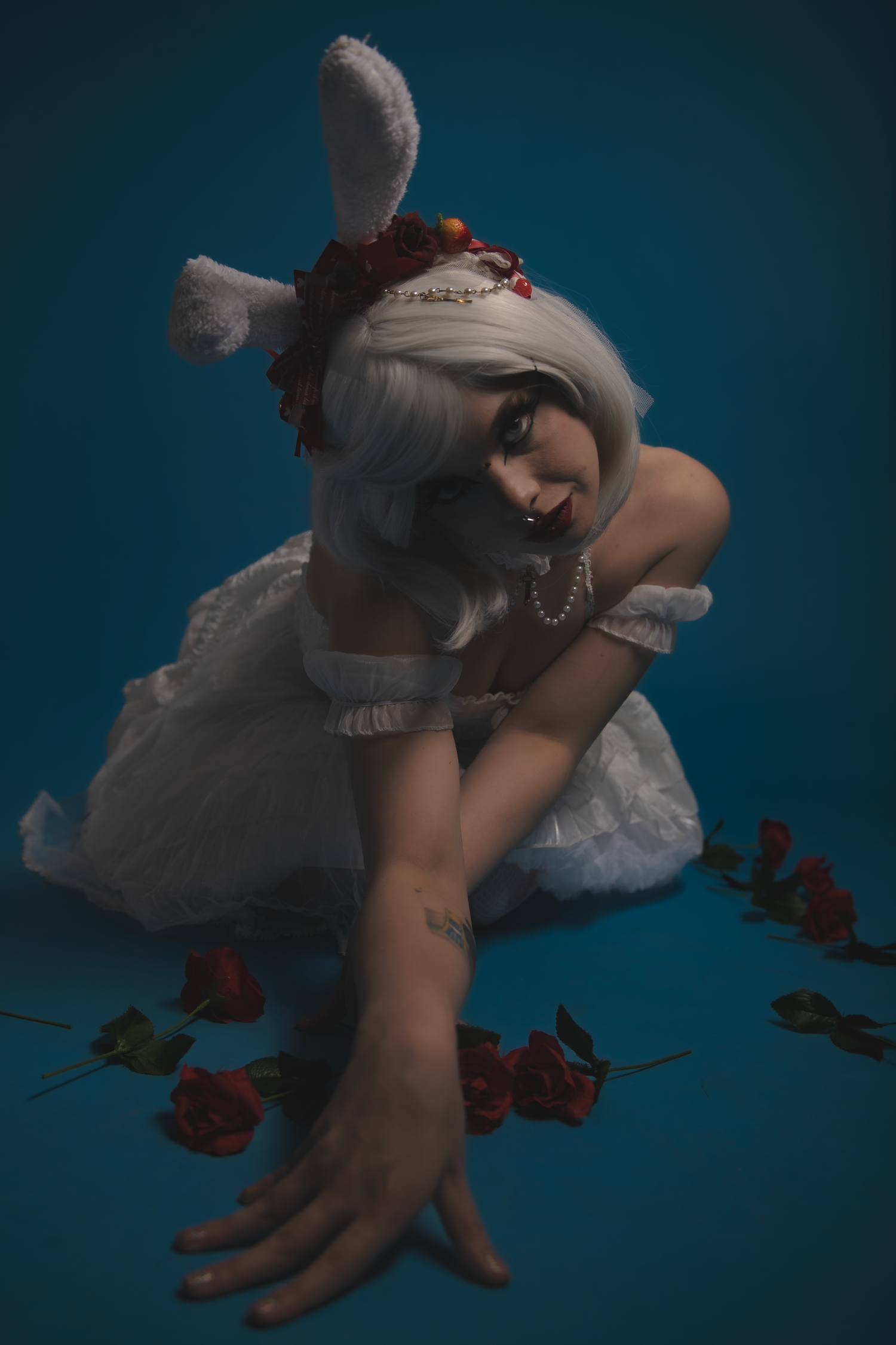 Moody artistic portrait featuring model in white dress with bunny ears against dark blue background with scattered roses.