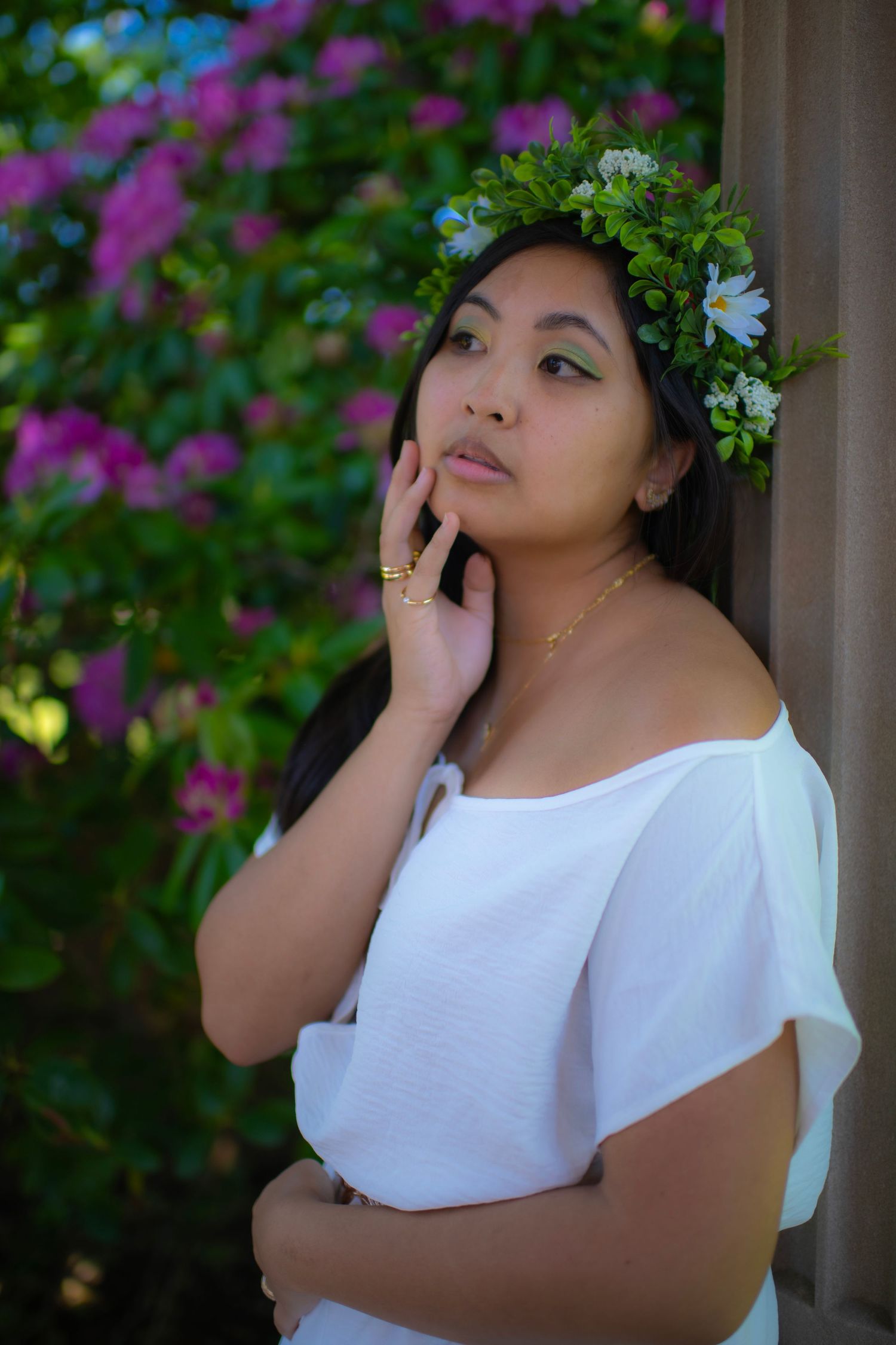 A thoughtful portrait in front of purple flowers featuring a white off-shoulder dress and floral crown of green leaves.