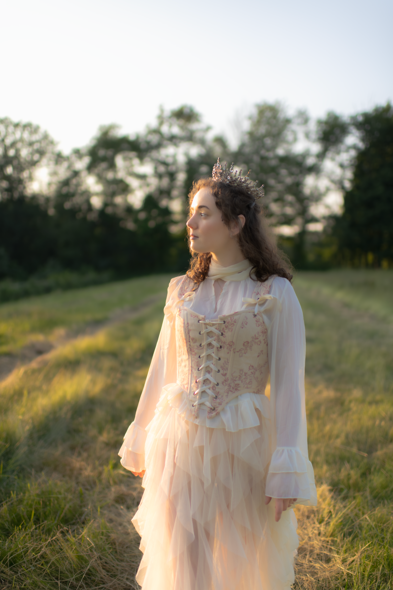 A person in a romantic white corset dress stands in a sunlit meadow at dusk.