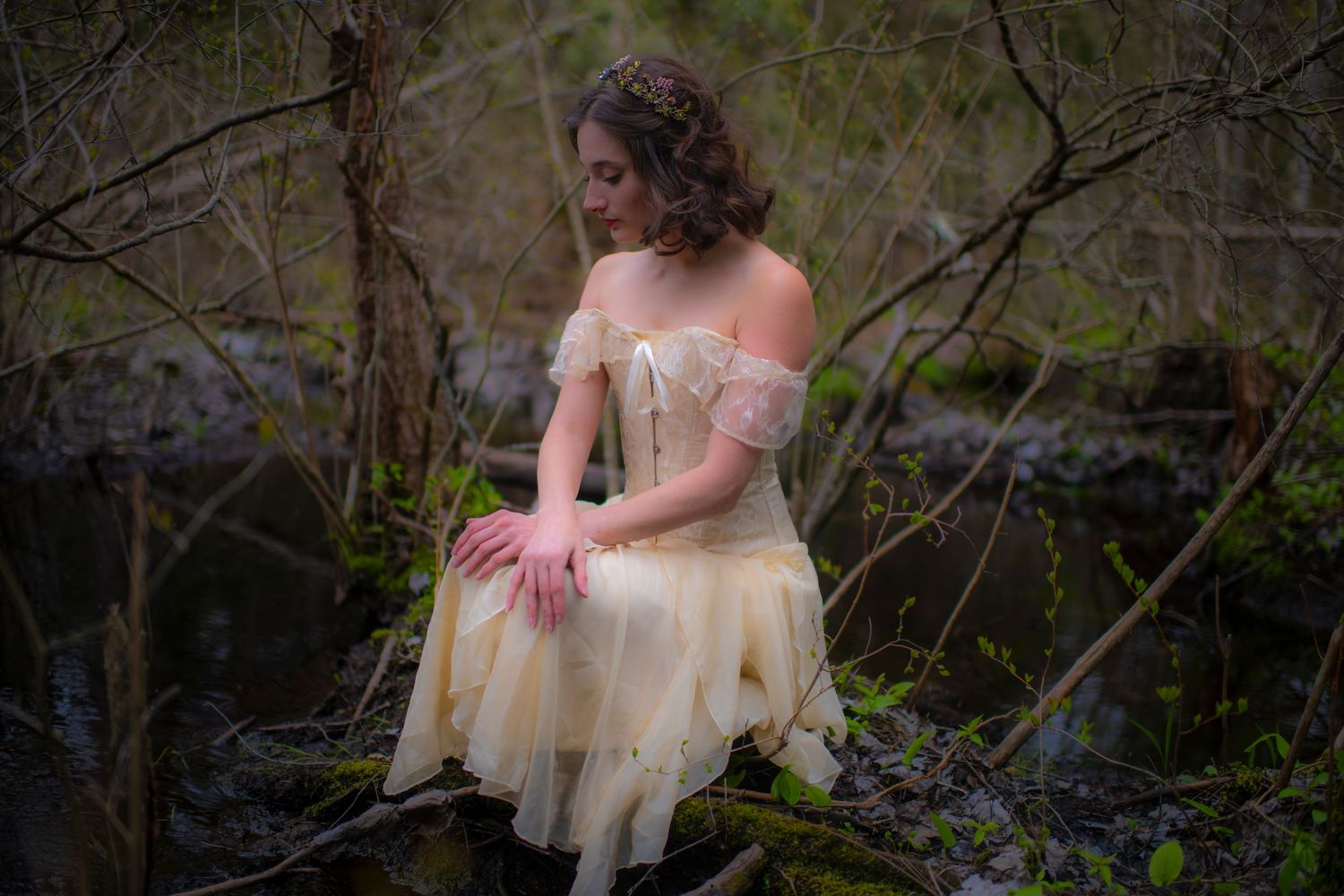 Figure in vintage tulle dress poses among branches by woodland pond.