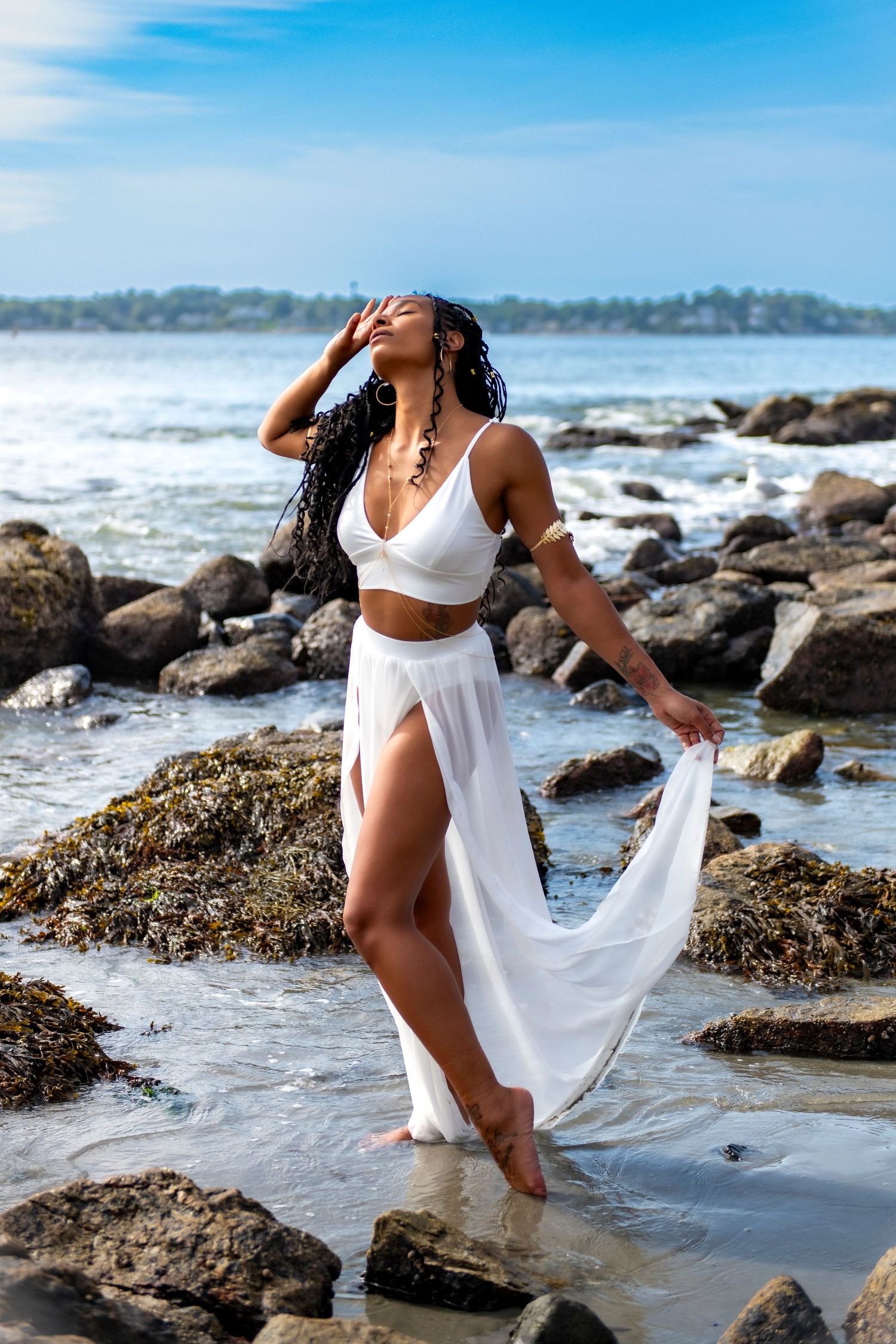 Woman in white beachwear poses dramatically on rocky coastline.