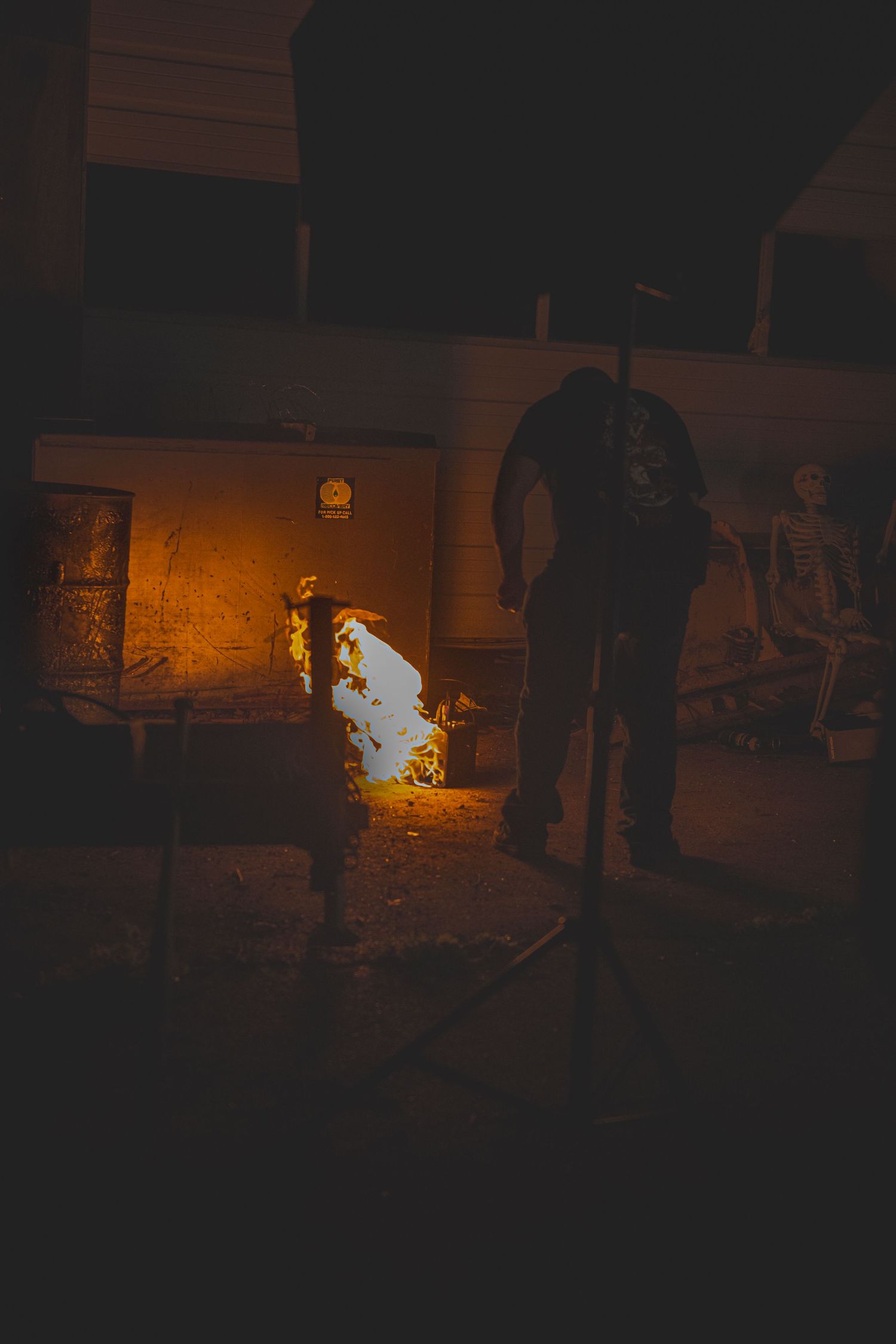 A silhouetted figure stands in the darkness near a small fire glowing orange against stone walls.