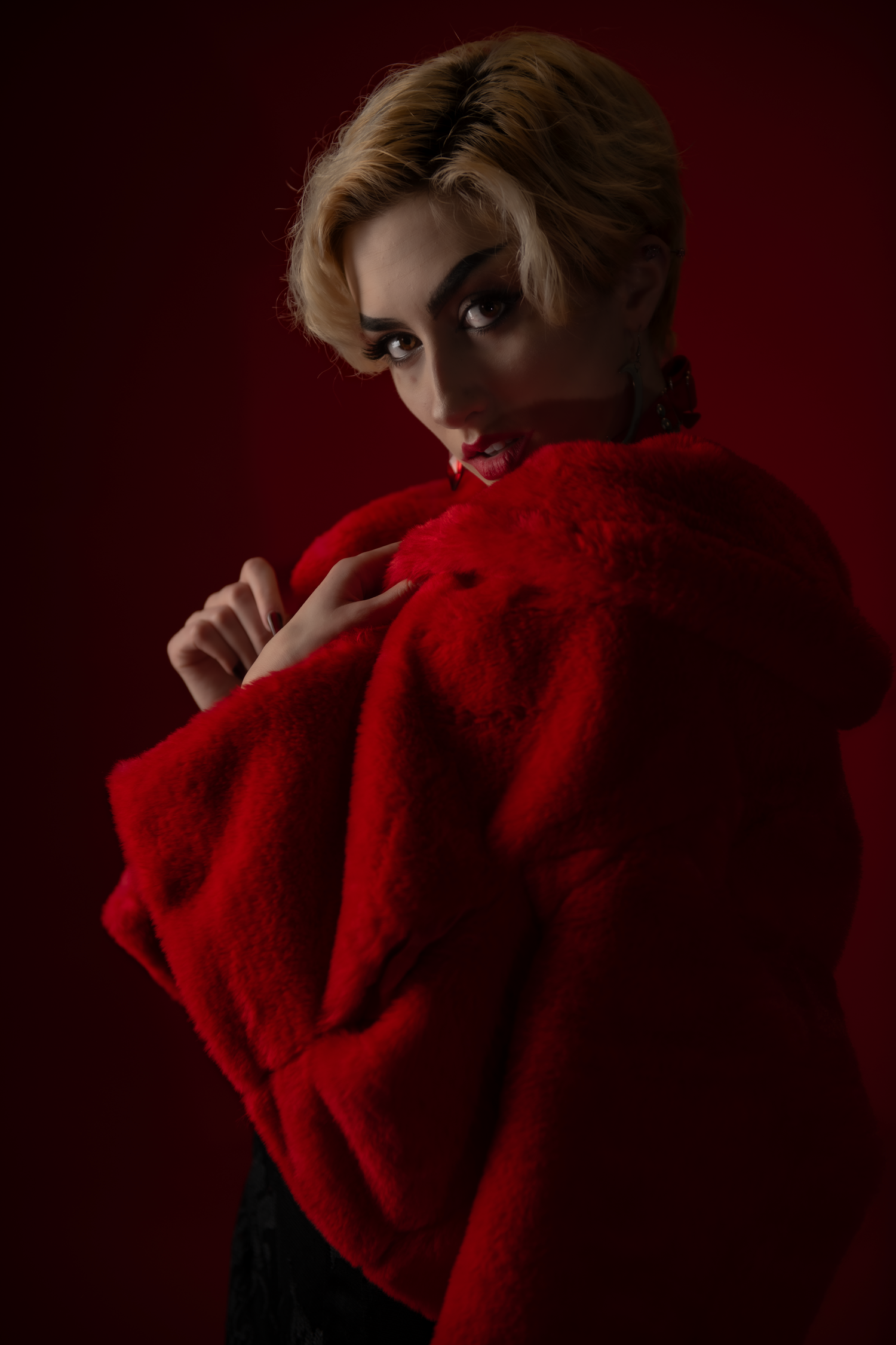 Dramatic portrait against dark red background features model in bright red fur coat with blonde hair in moody lighting.