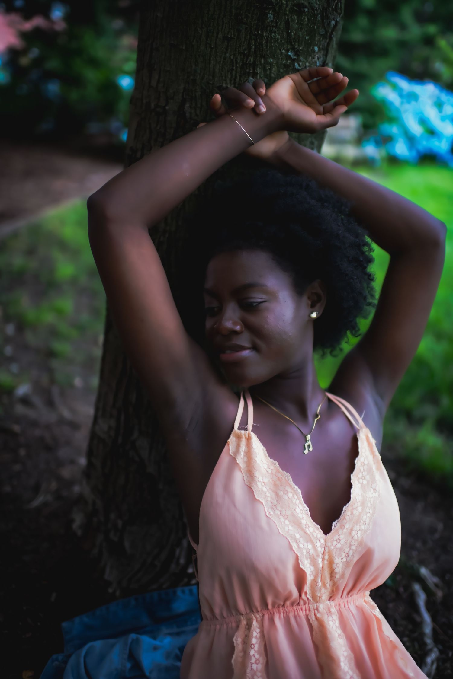 Graceful figure in peach dress poses against a tree trunk in a natural outdoor setting.