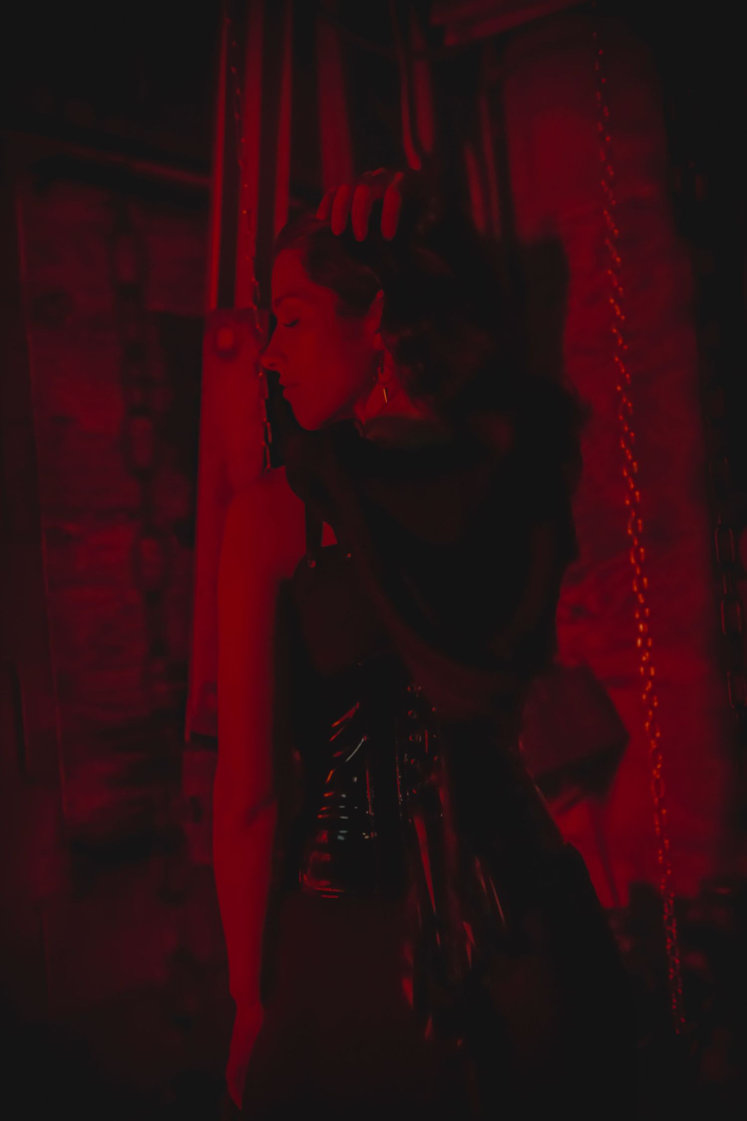 Dramatic silhouette figure in gothic attire against red backlit background with hanging chains creating dark mysterious ambiance.