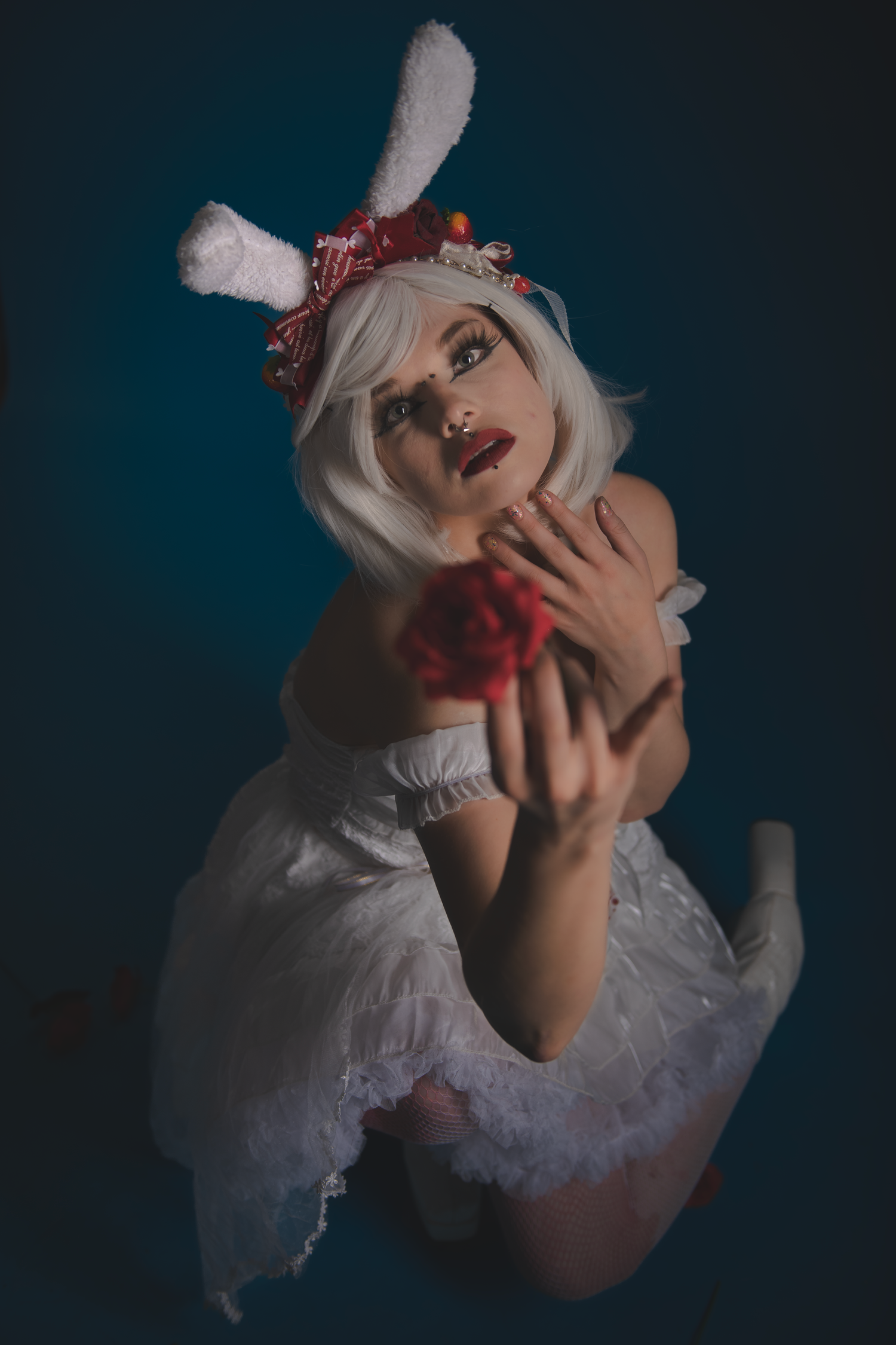 Dramatic portrait in dark lighting features figure in white dress holding red flower.