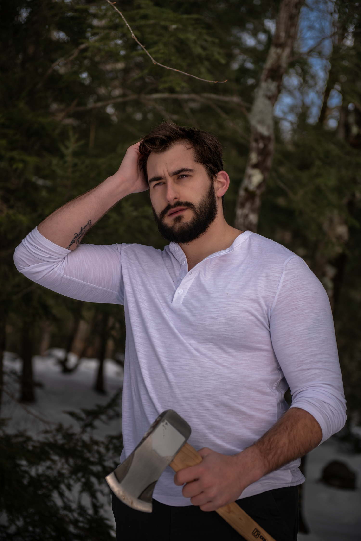 Man in white henley shirt holds an axe while standing in snowy forest setting at dusk.