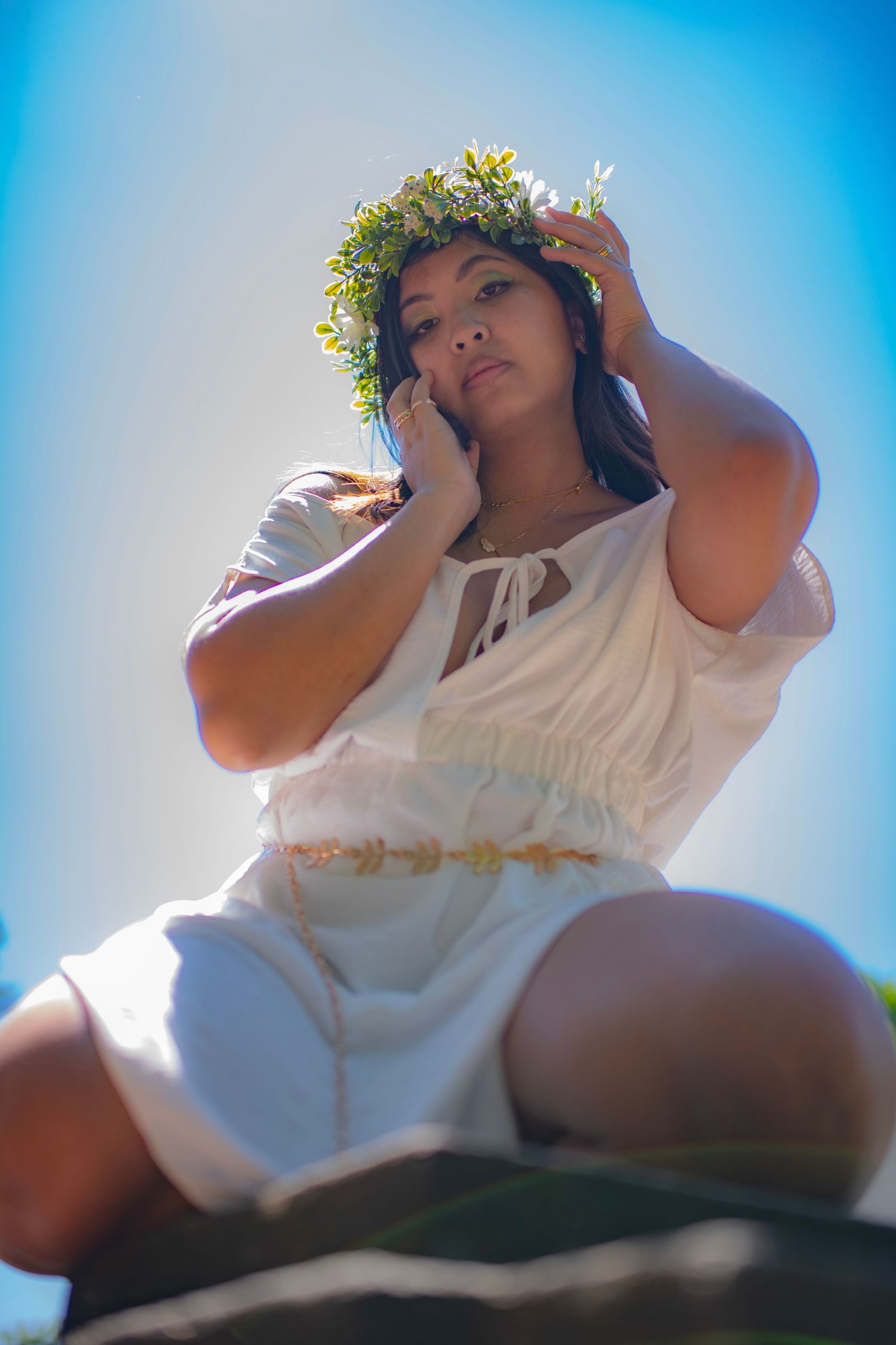 A person in a flowing white dress and floral crown poses outdoors against a bright blue sky in dreamy sunlight.
