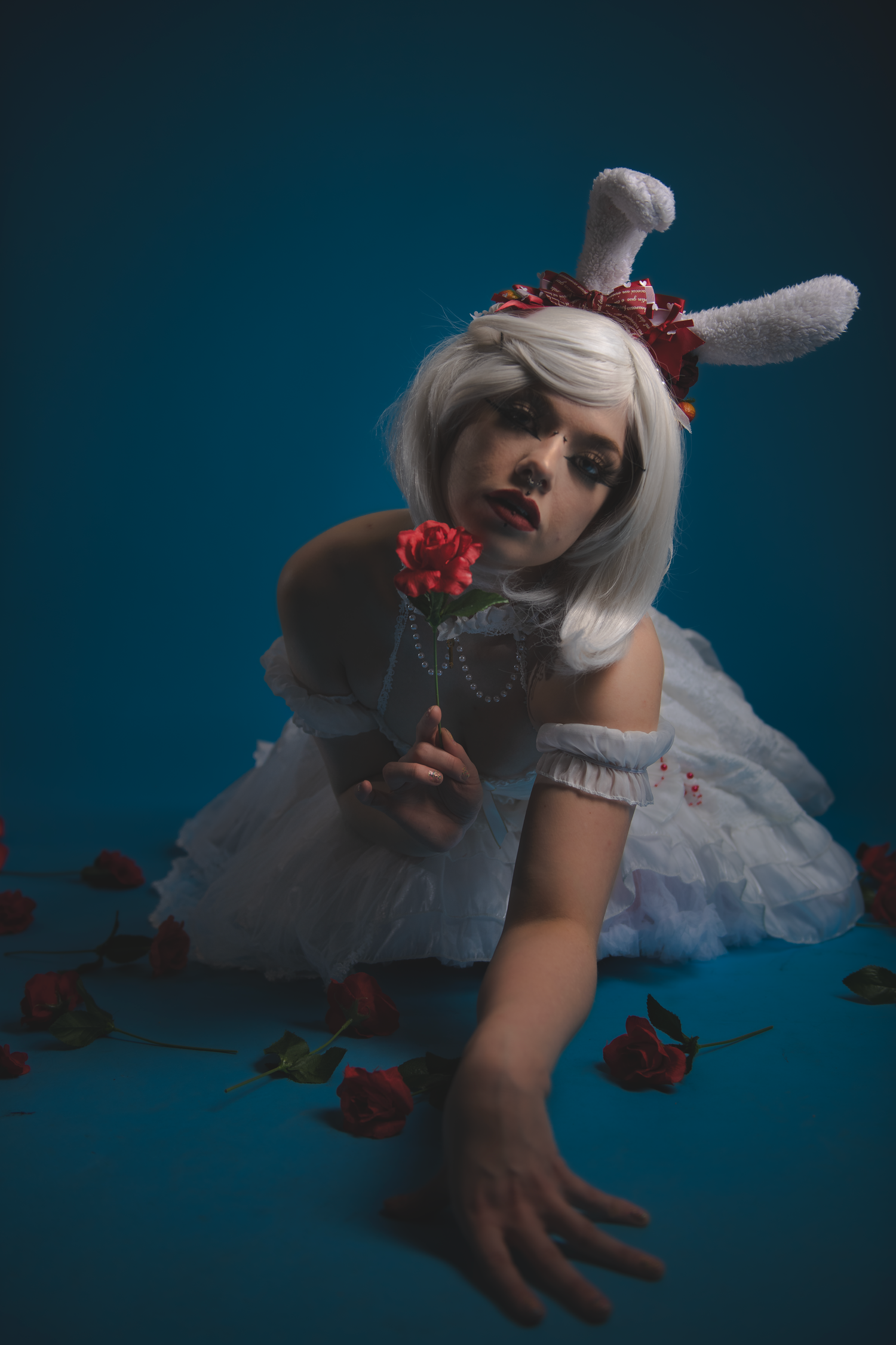 A whimsical figure in white dress and bunny ears lies on the floor holding a red rose against a dark teal backdrop.