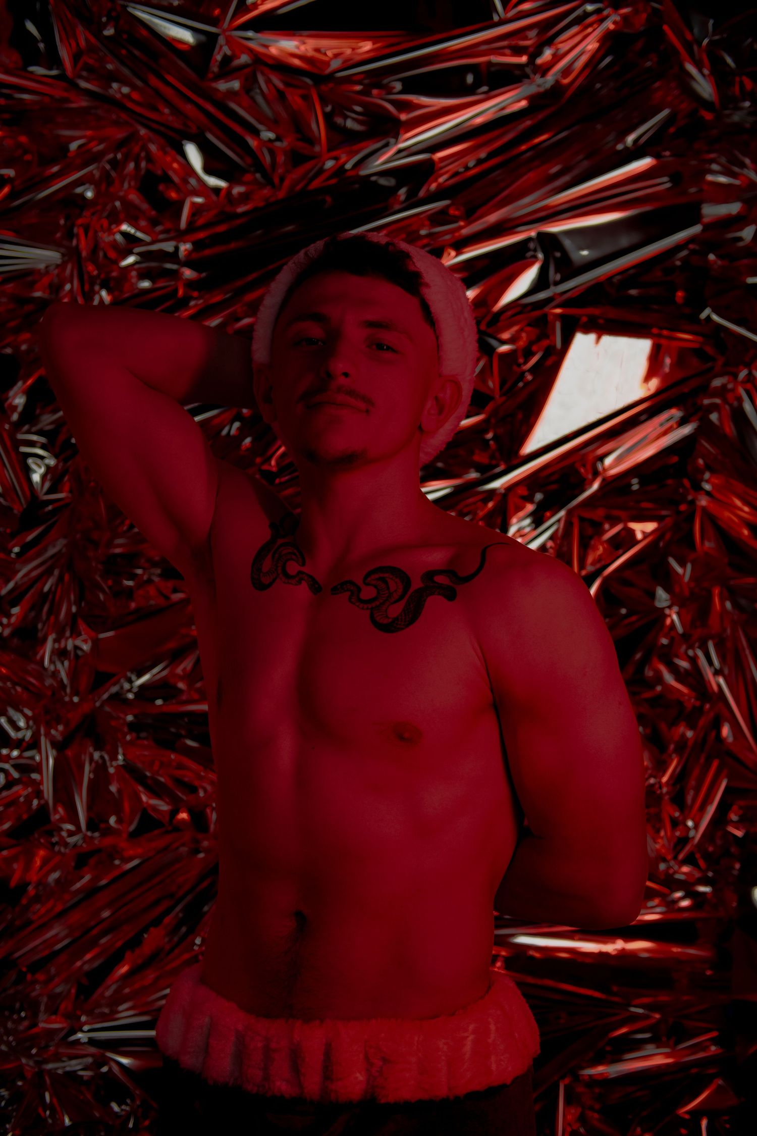 Shirtless figure posed against dramatic red metallic background with reflective chrome-like surfaces in moody lighting.