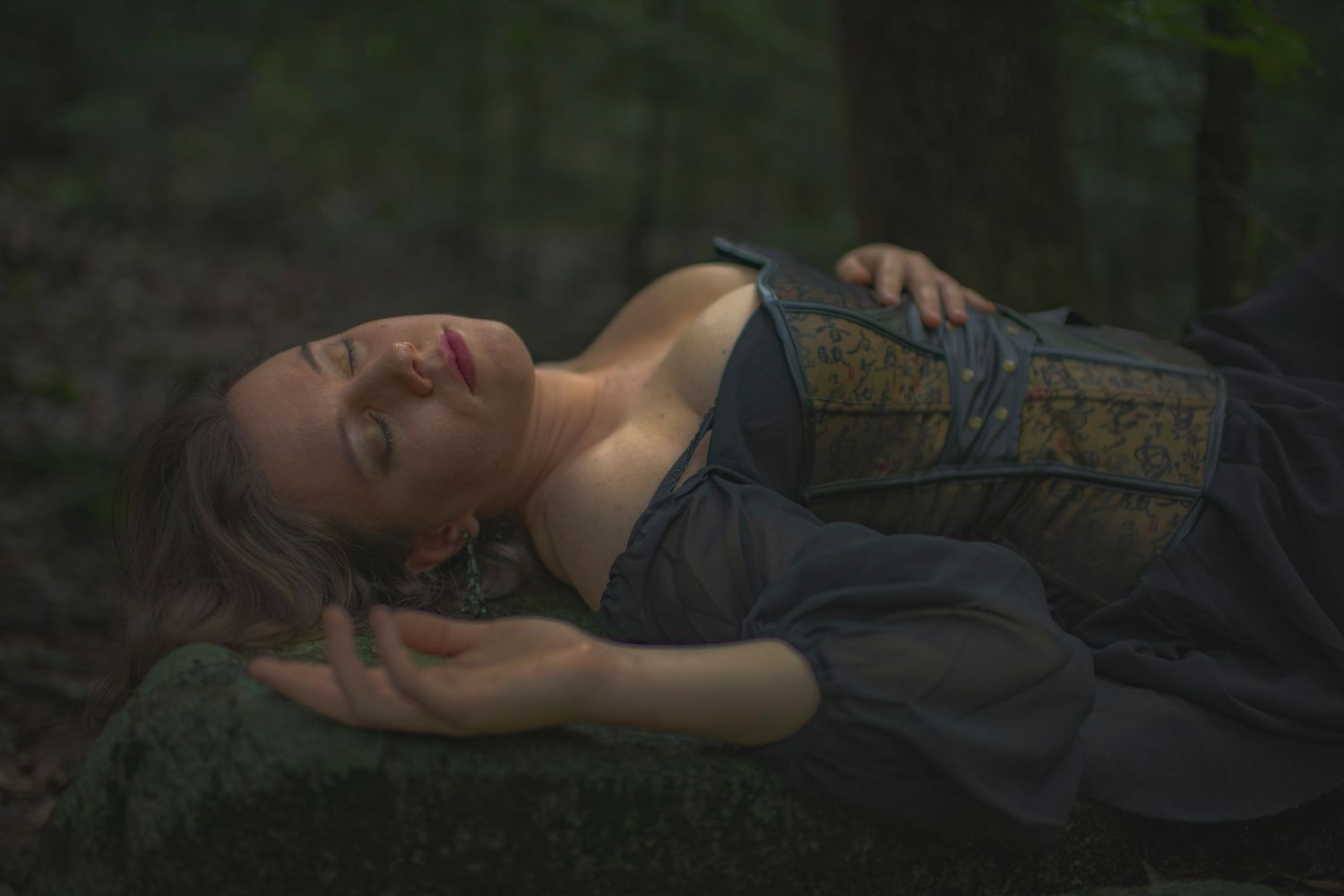Atmospheric forest photo of someone reclining on moss-covered ground in period costume.