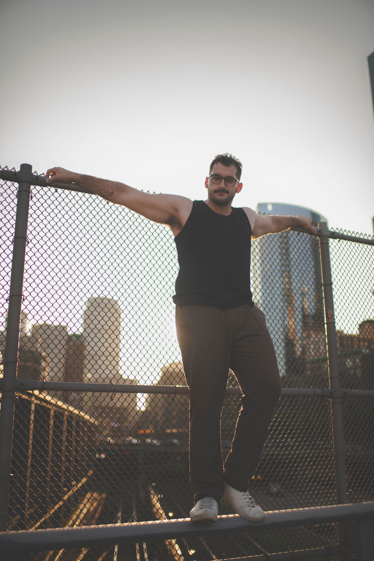 Person in black attire poses on chain link fence with city skyline at sunset creating dramatic silhouette.