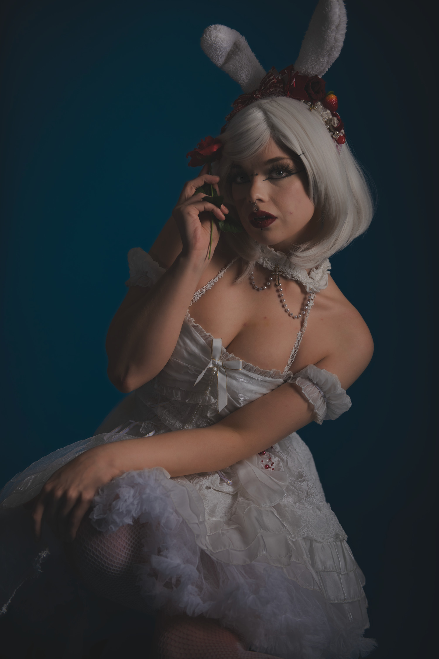 Figure in white lace dress and bunny accessories posing dramatically on dark backdrop.