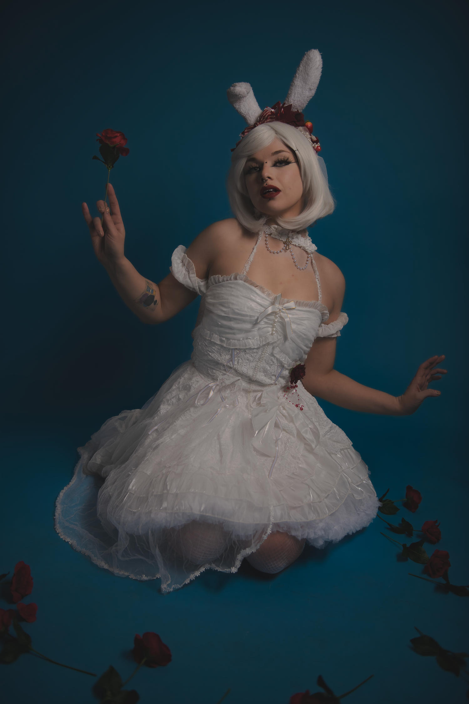 A figure in a white tulle dress and bunny ears poses gracefully among floating red roses against a dark teal backdrop.