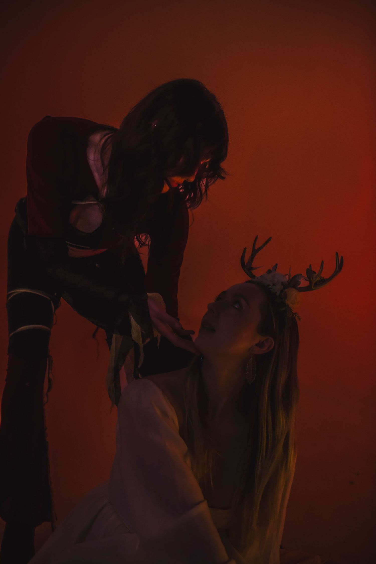 Dark artistic photo of two figures in dramatic poses against a red background, with one wearing antlers.