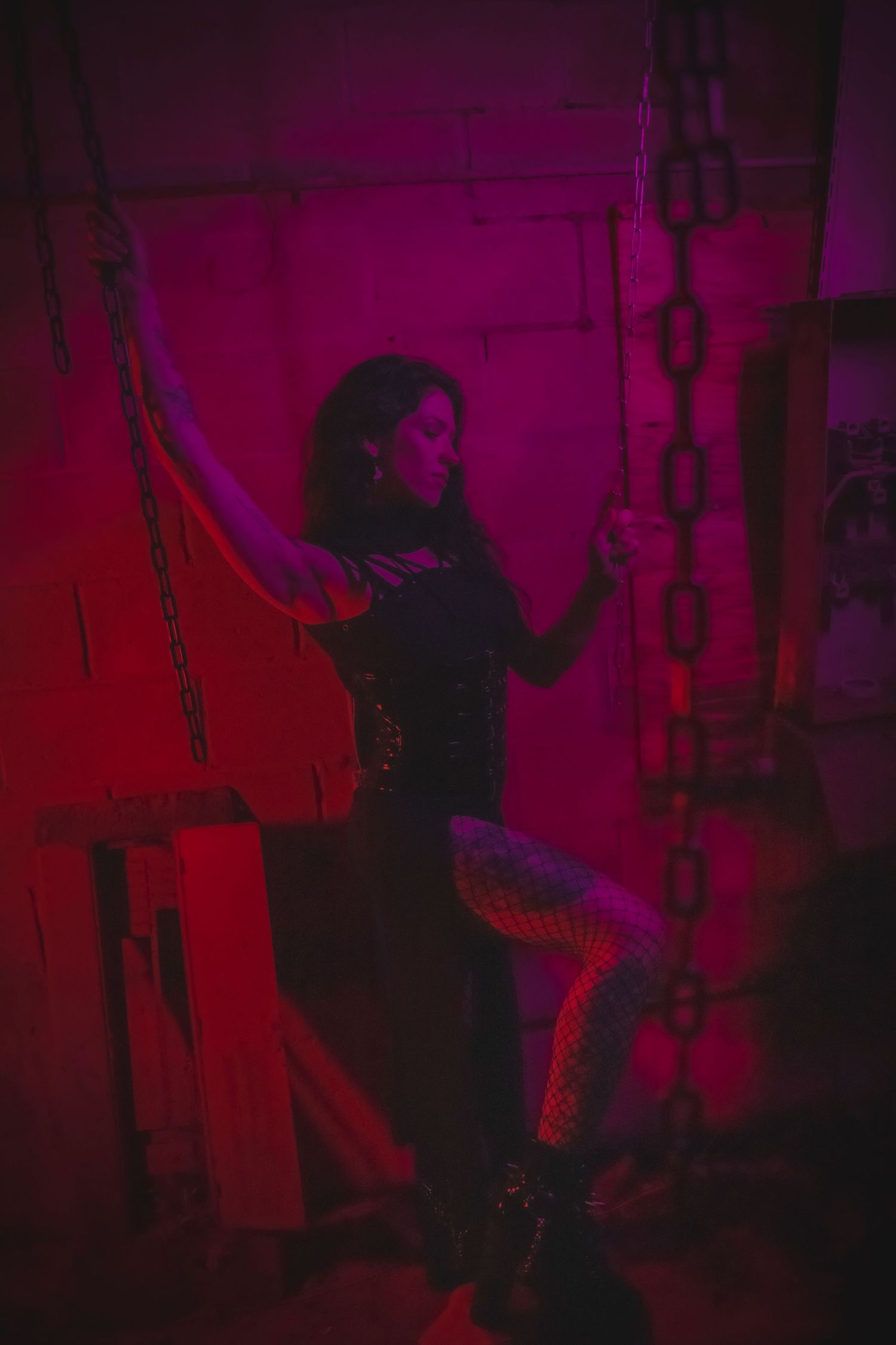 Silhouette of figure in black outfit stands against red-lit wall with chains, creating a moody atmosphere.
