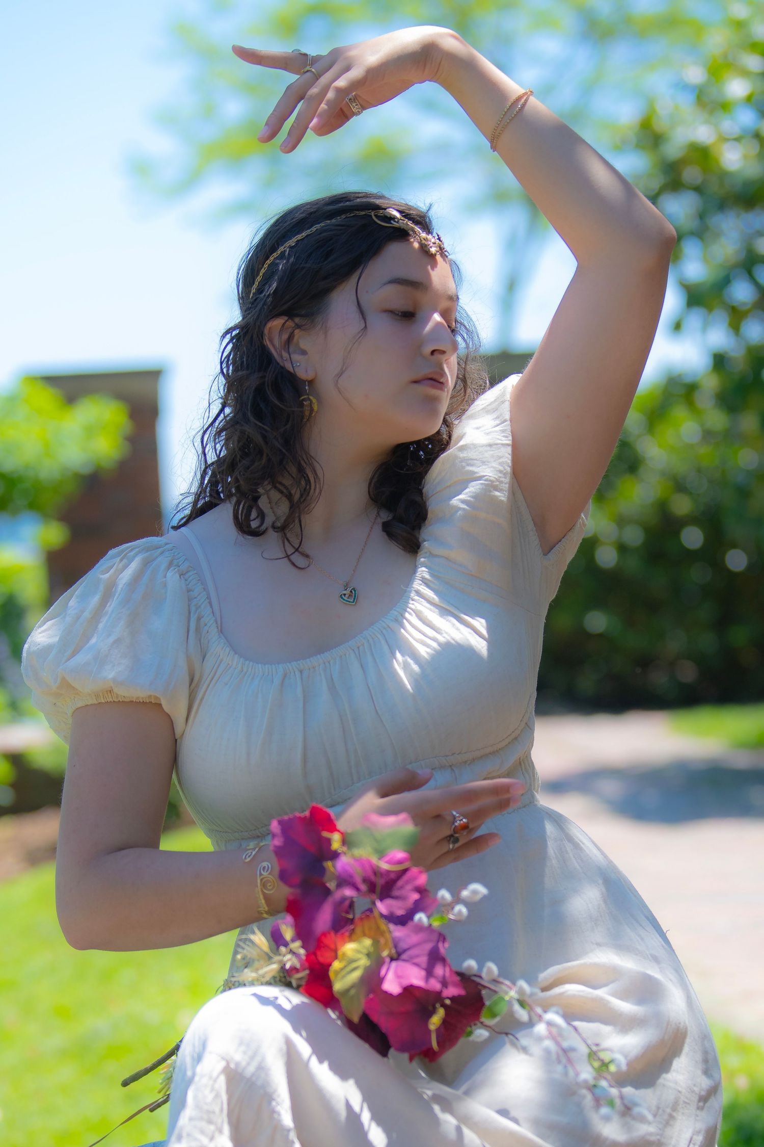 Person in white dress poses gracefully outdoors with colorful flowers on sunny day among green foliage.