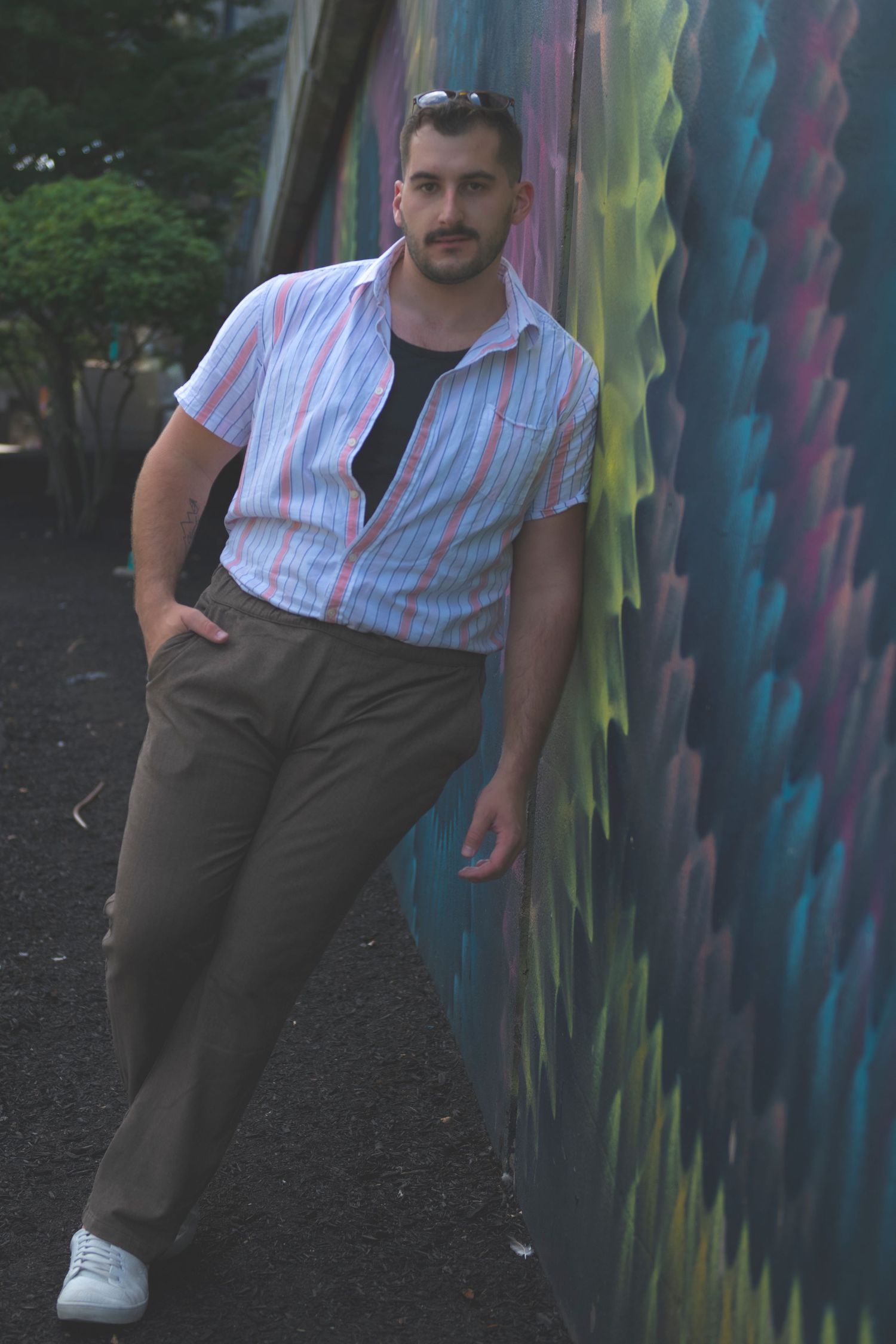 A person wearing a striped button-down shirt and dark pants poses against a colorful graffiti wall outdoors.