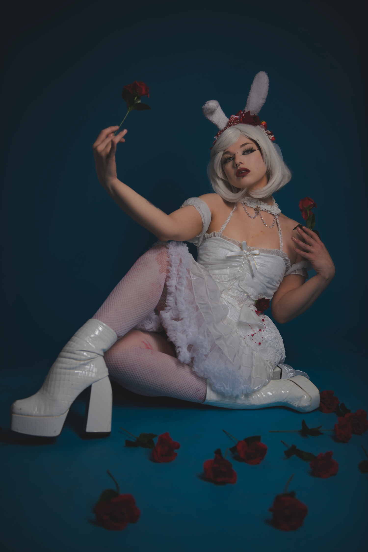 Figure in white frilly dress and bunny ears poses with red roses on dark blue background.