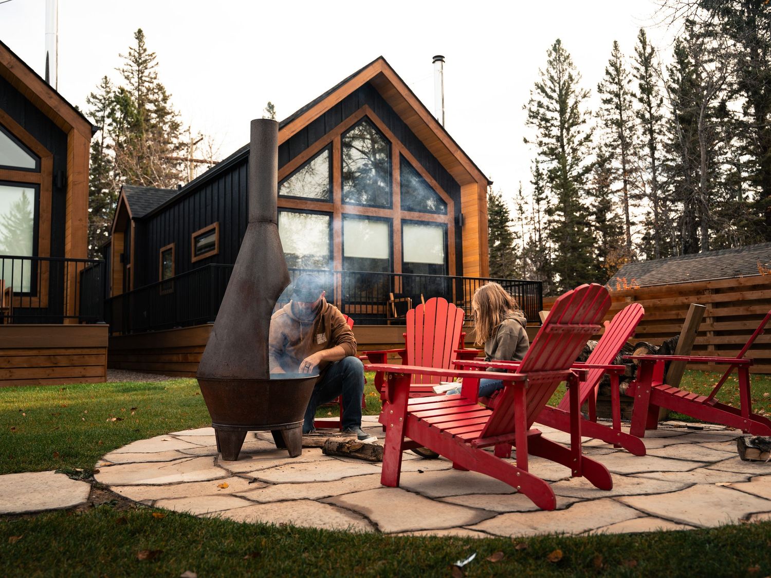 Modern cabin with large windows and red Adirondack chairs around a fire pit.