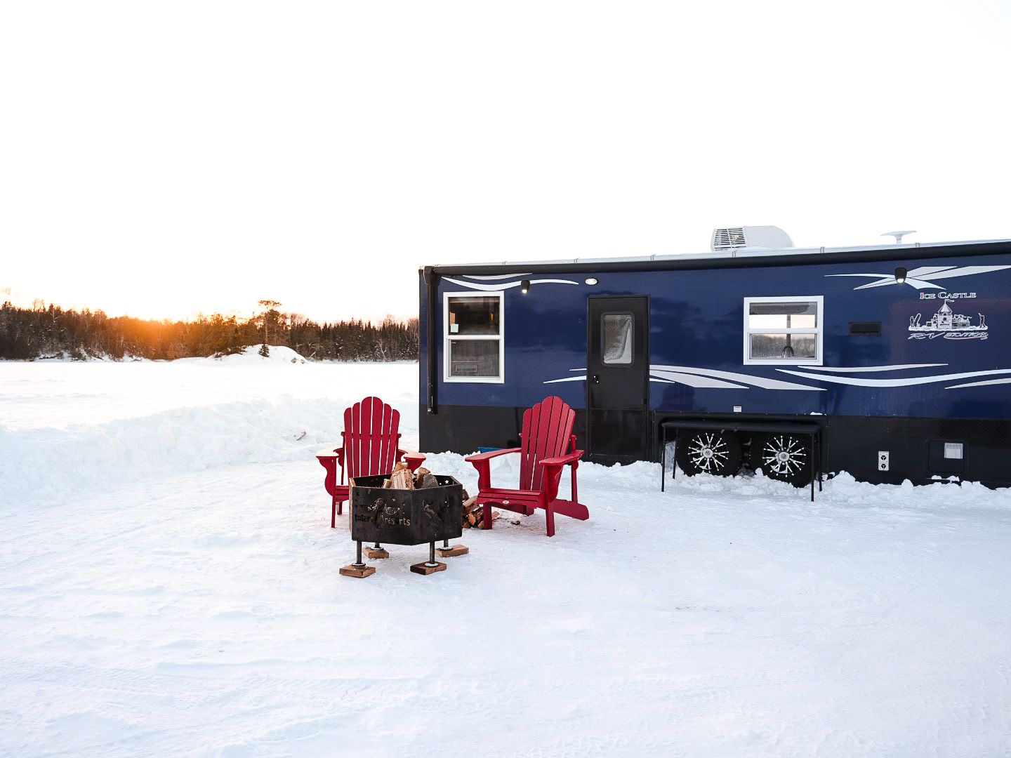 Snow-covered landscape with blue cabins and red Adirondack chairs near a frozen lake.