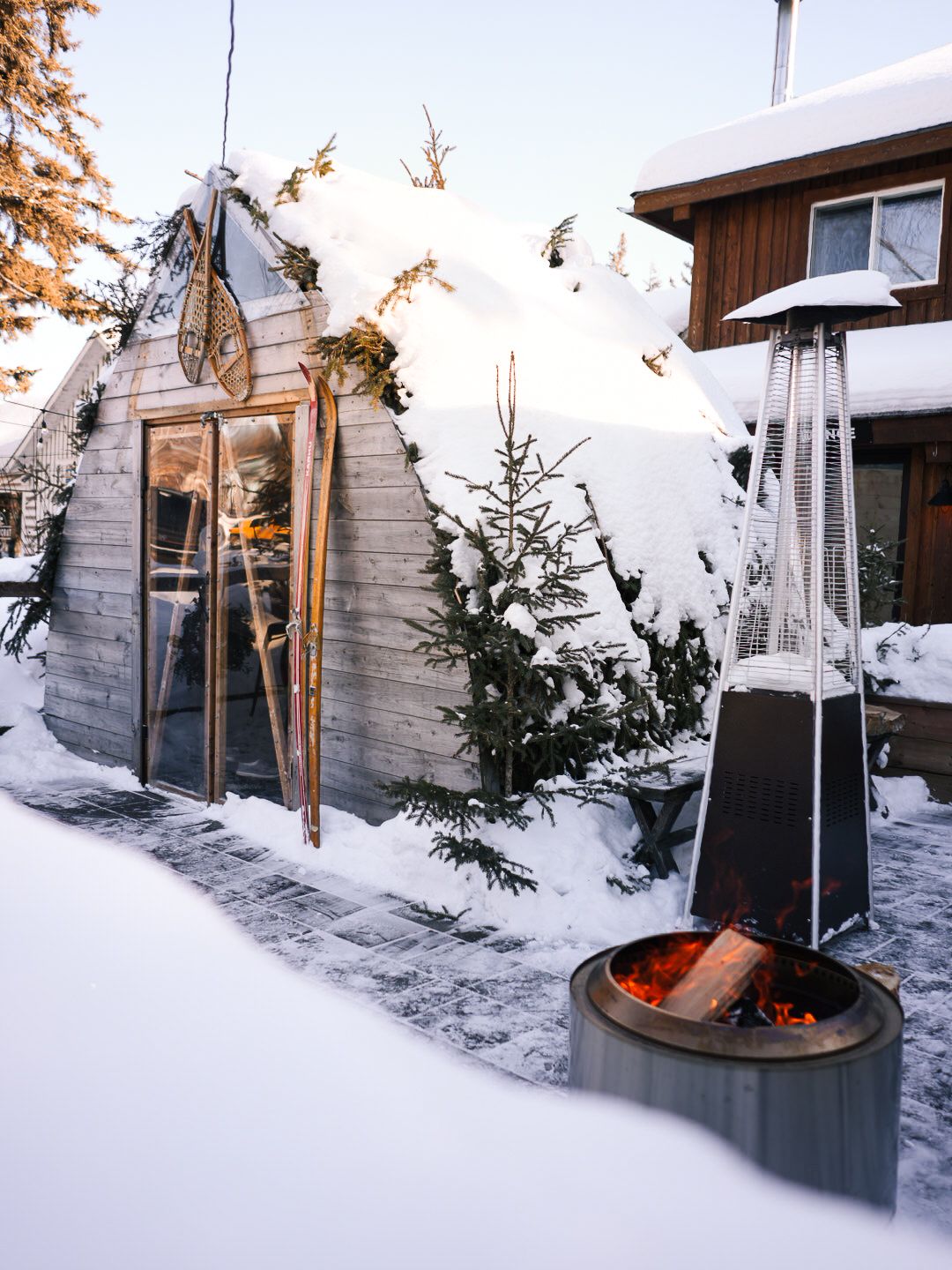 Modern outdoor fire pit surrounded by snow with wooden cabin in background during sunset.