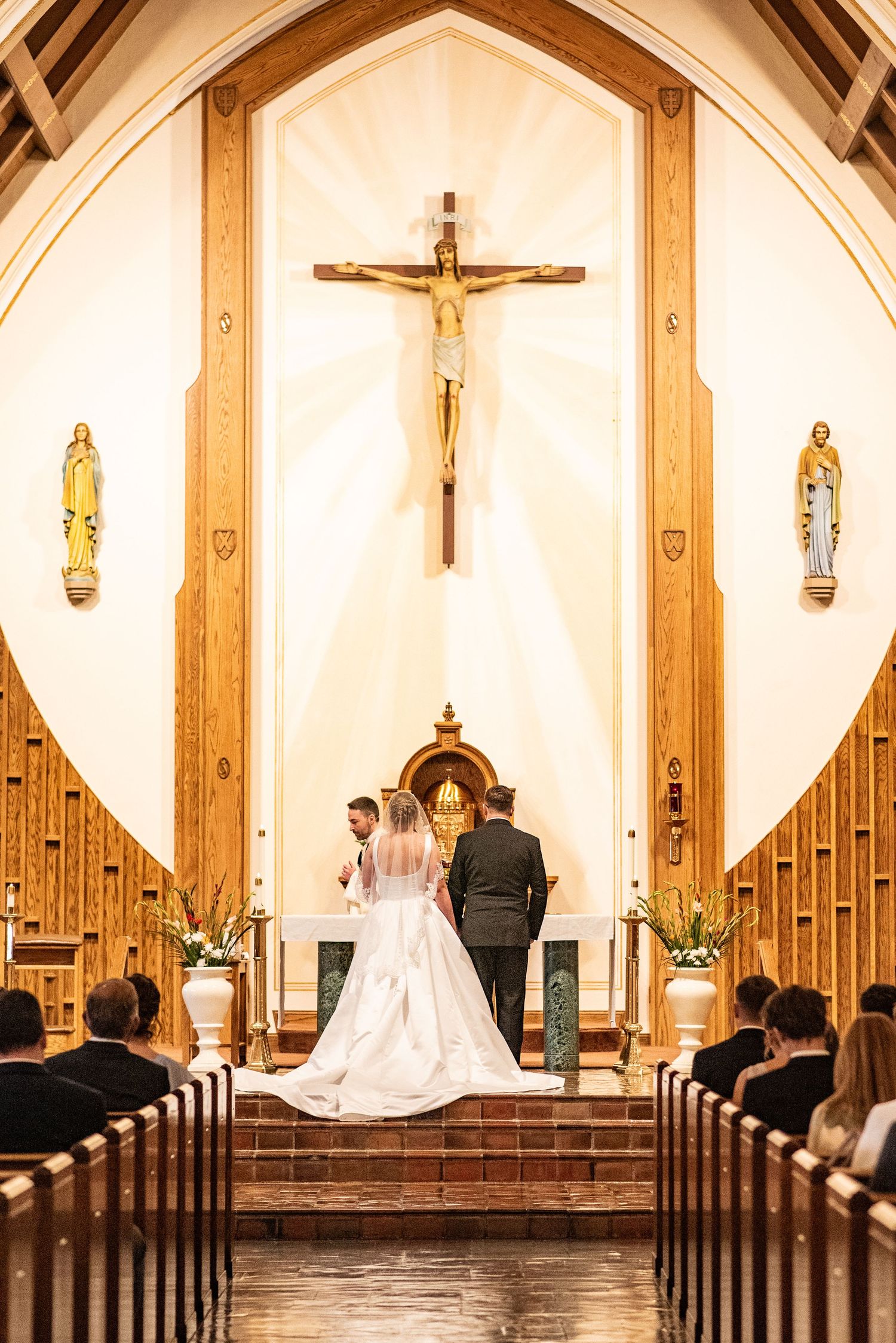 Bride and groom exchange vows at Catholic church altar beneath crucifix, guests seated in wooden pews.