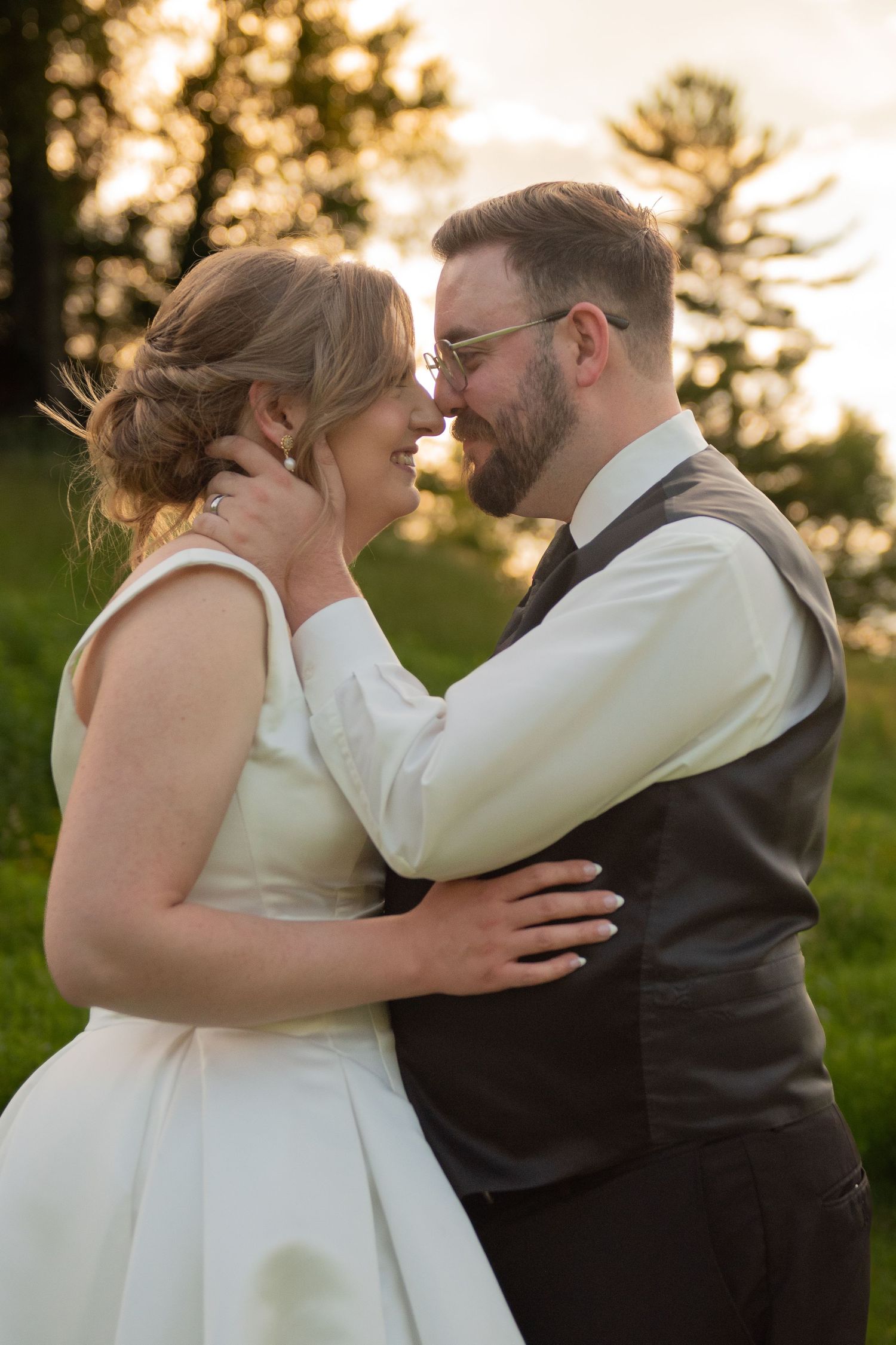A bride and groom share a romantic moment at sunset, embracing and smiling at each other in a lush green outdoor setting.