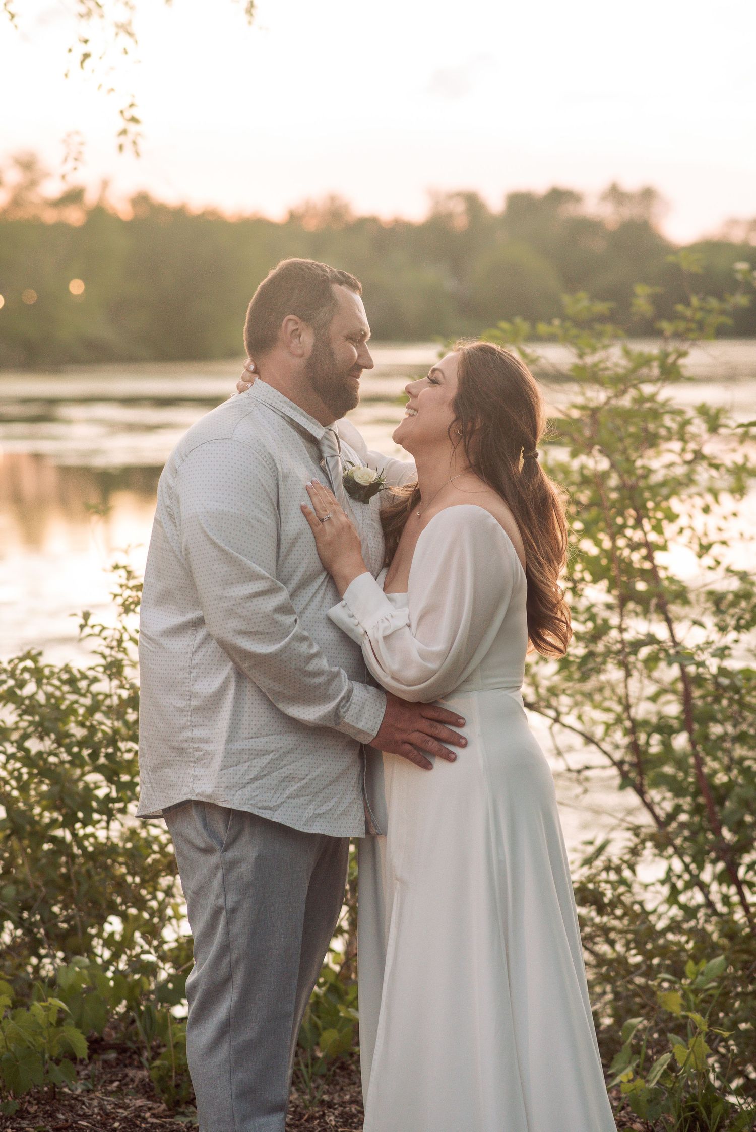 Couple in wedding attire embracing at golden hour by a serene lake, bride in white gown, groom in gray suit.