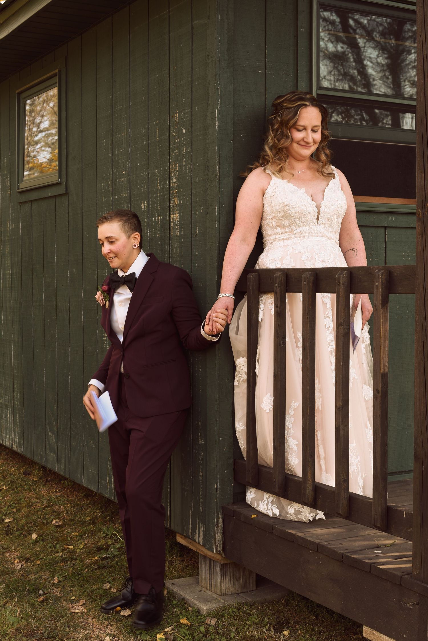 Two brides hold hands on rustic green cabin steps, one in a white lace gown, one in a burgundy suit.
