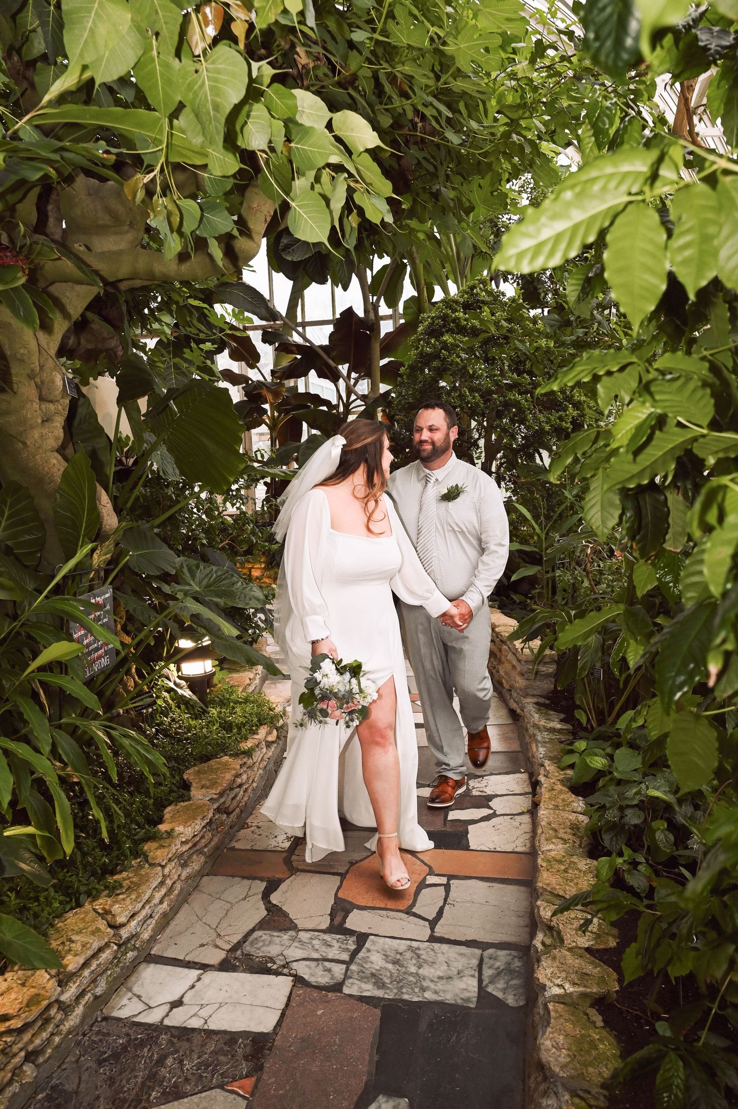 Bride and groom walk a lush tropical garden stone path, bride in white dress holding bouquet, groom in gray suit smiling.