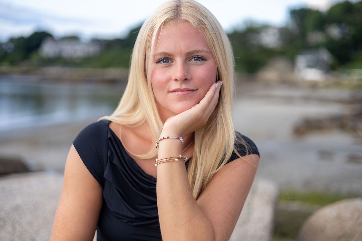 A person poses outdoors near a waterfront setting wearing a black top and long blonde hair.