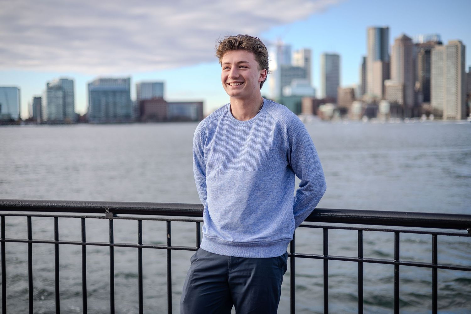 Person in light blue sweater leans against railing with Boston harbor and city skyline in background.