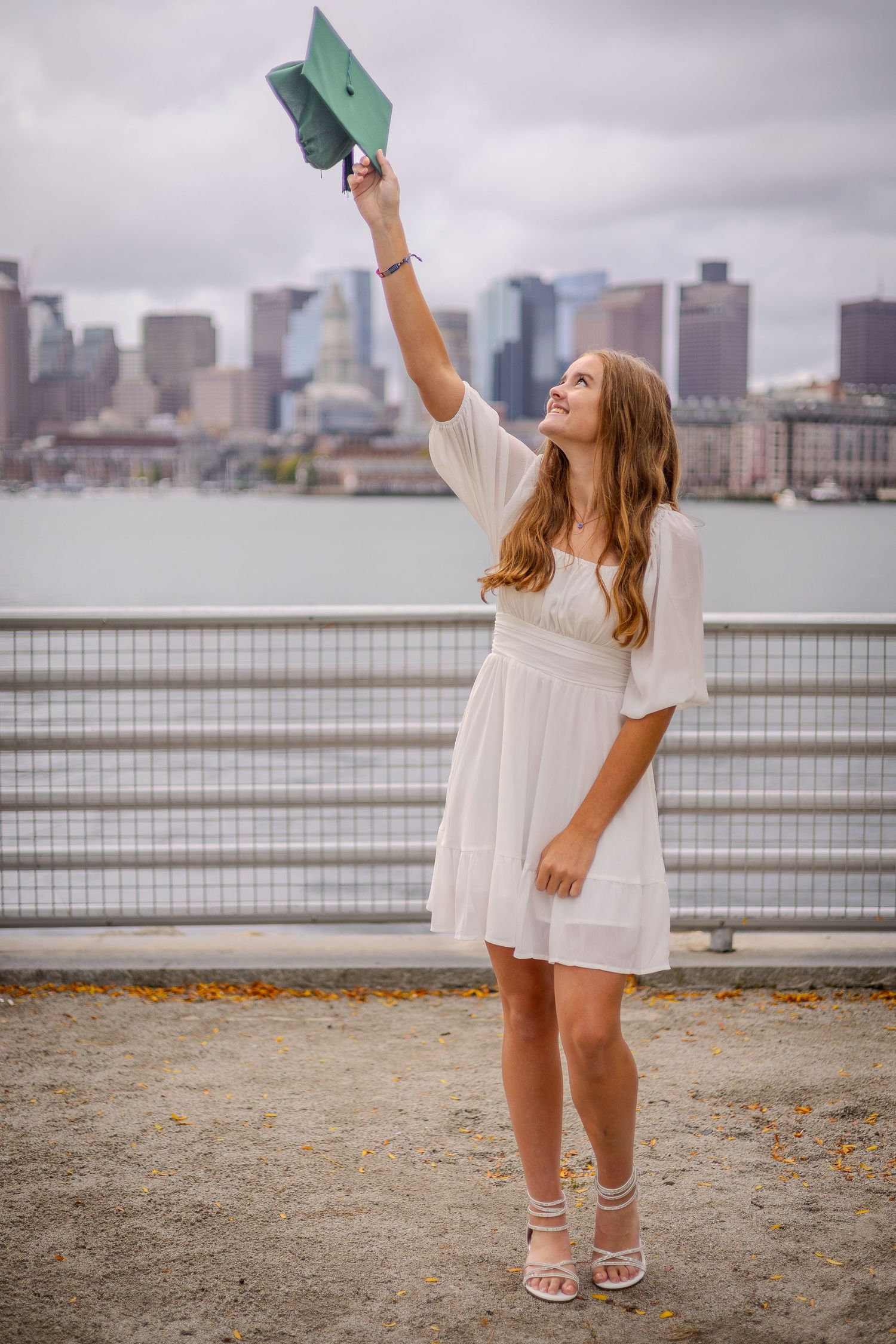 Graduate in white dress holds up diploma against urban skyline backdrop on overcast day.