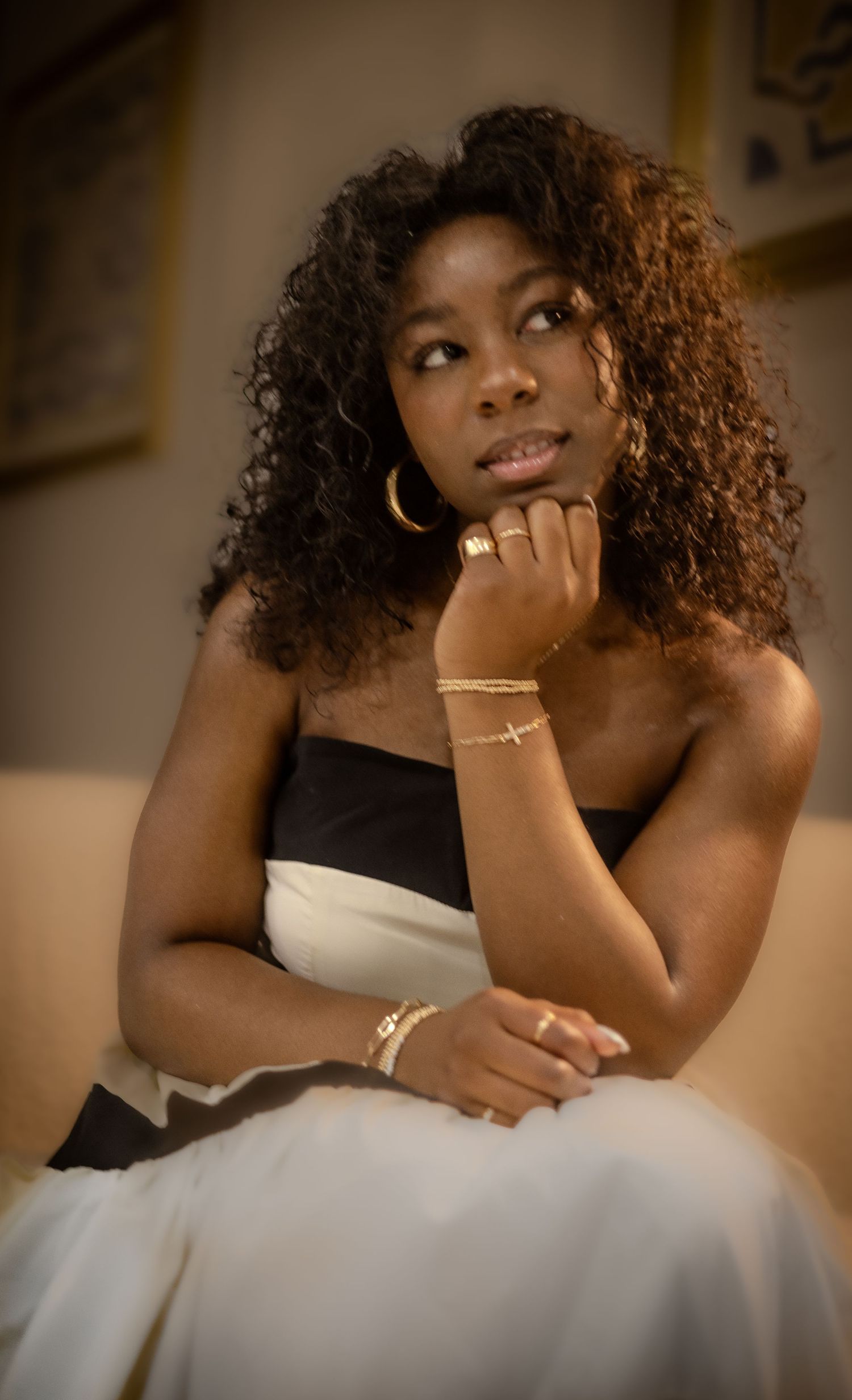 An elegant portrait featuring curly hair, gold jewelry, and a black and white dress in warm lighting.