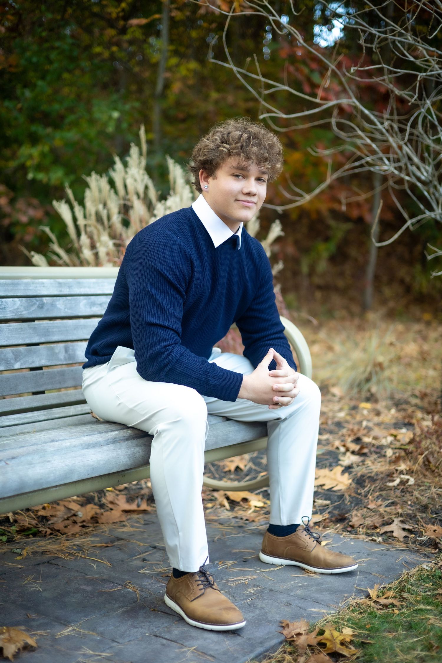 A person wearing a navy blue sweater and white pants sits thoughtfully on a wooden bench surrounded by fallen autumn leaves.