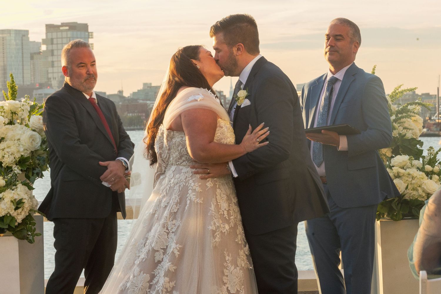 A couple shares a romantic moment during their outdoor wedding ceremony with guests looking on at sunset.